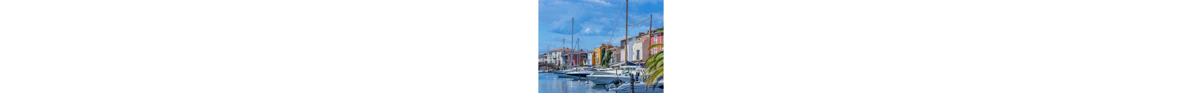 A row of sleek motorboats and sailboats is docked along a calm waterfront marina. Colorful multi-story buildings line the quay, reflecting in the water under a bright blue sky with scattered clouds. A palm tree frames the right side of the scene, adding a Mediterranean coastal feel. The overall atmosphere is peaceful and leisurely, suggesting a relaxed seaside town or vacation destination.
