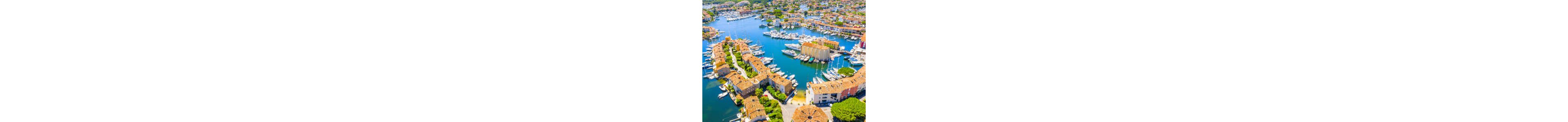 An aerial view captures a picturesque Mediterranean coastal town built around a calm marina. Rows of terracotta-roofed houses line narrow canals filled with numerous white yachts and sailboats. The bright blue water contrasts with the warm earth-toned buildings and lush green trees scattered throughout the scene. In the distance, more residential areas and boats extend toward the horizon, emphasizing a vibrant seaside lifestyle.