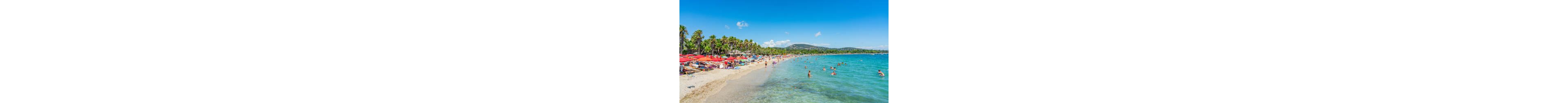 A lively sandy beach is filled with people relaxing under colorful umbrellas and sun loungers. Swimmers and waders enjoy the clear turquoise sea close to the shoreline. Palm trees line the beach, adding a tropical feel to the scene. Green hills and a bright blue sky with no visible clouds create a warm, sunny holiday atmosphere.