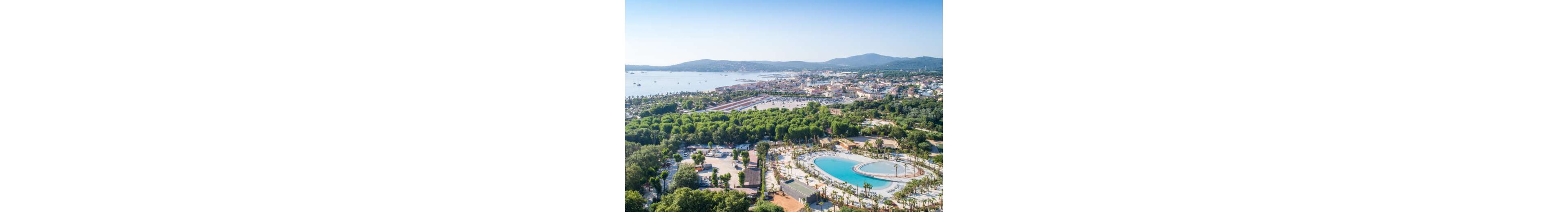 An aerial view showcases a large coastal resort complex with a prominent circular lagoon-style pool in the foreground. The resort is surrounded by dense green trees and landscaped pathways, creating a lush environment. Beyond the resort, a bay with calm water and a distant shoreline town stretches across the horizon. Rolling hills frame the background, adding depth to the scenic coastal landscape.