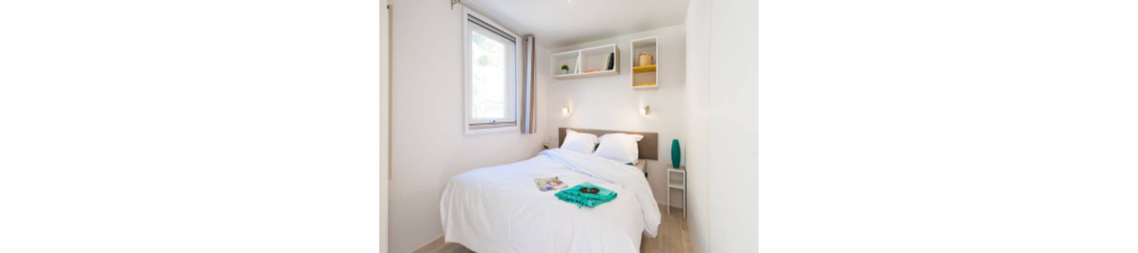 A small modern bedroom features a neatly made double bed with crisp white bedding. The room is bright, with natural light coming through a single window on the left. Open wall shelves above the headboard display a few decorative items, while a small bedside table holds a turquoise vase. A folded turquoise towel and a magazine rest on the bed, adding a touch of color to the otherwise neutral space.