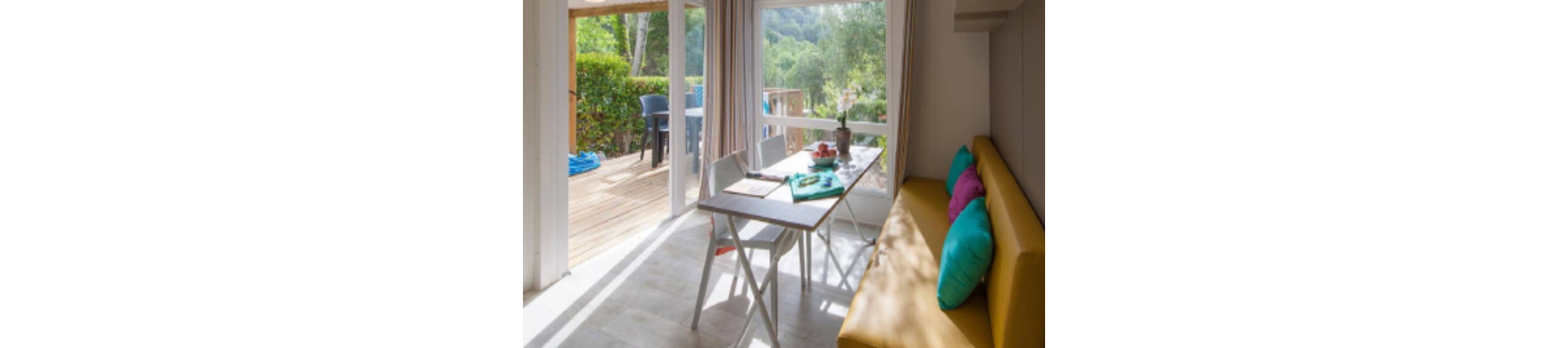 A modern holiday cabin dining area features a slim table with chairs and a long yellow bench lined with colorful cushions. Large windows and a glass door open onto a wooden terrace with outdoor seating. Sunlight fills the room, highlighting light flooring and simple decor, including a small plant on the table. Lush greenery and trees are visible outside, creating a relaxed vacation atmosphere.