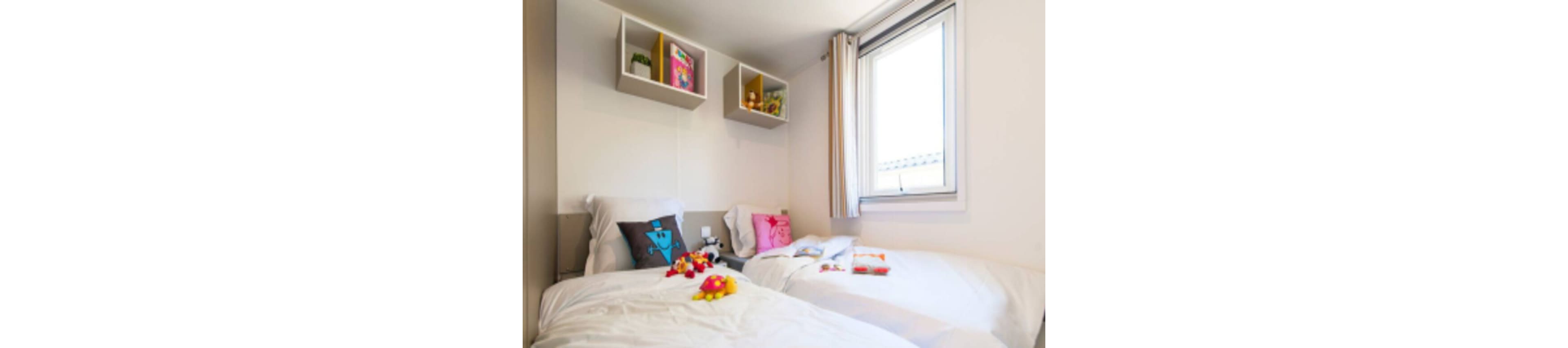 A small children's bedroom features two neatly made twin beds with white bedding. Colorful plush toys and character cushions are placed on the beds, adding a playful touch. Wall-mounted shelves above the headboards display books, toys, and decorative items. Natural light enters through a window with simple curtains, creating a bright and cozy atmosphere.