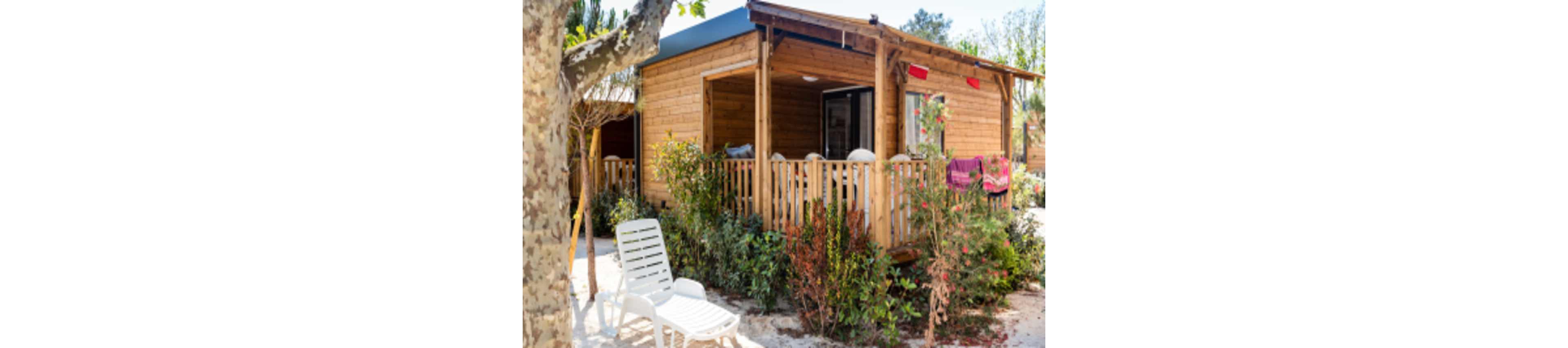 A small wooden cabin with a covered porch is surrounded by lush greenery and flowering shrubs. White plastic lounge chairs sit in the sun near a tree in the foreground. Colorful towels hang over the porch railing, suggesting a relaxed holiday atmosphere. The scene is bright and inviting, with clear daylight and a casual outdoor setting.