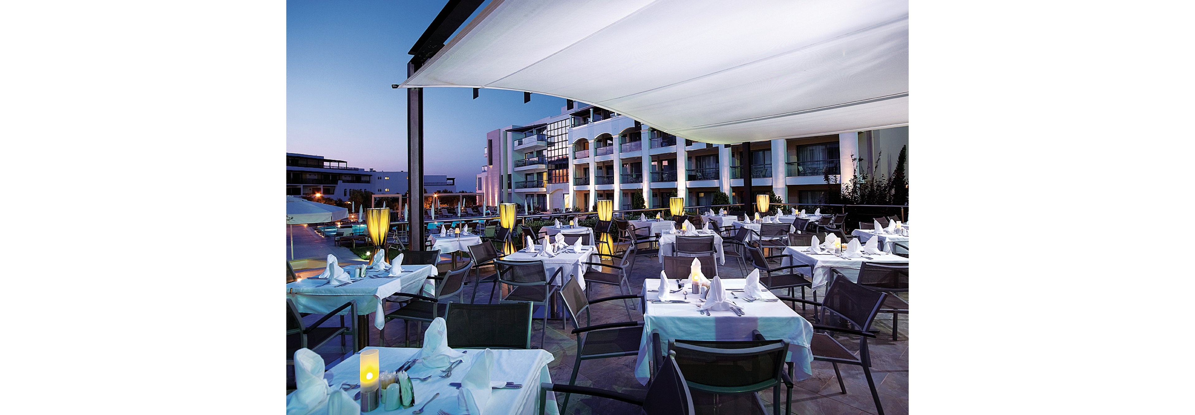 An elegant outdoor dining area set at a luxury resort during twilight. Tables are neatly arranged with white tablecloths and ambient lighting, creating a serene atmosphere. The backdrop features modern architecture and a poolside view, enhancing the upscale setting.