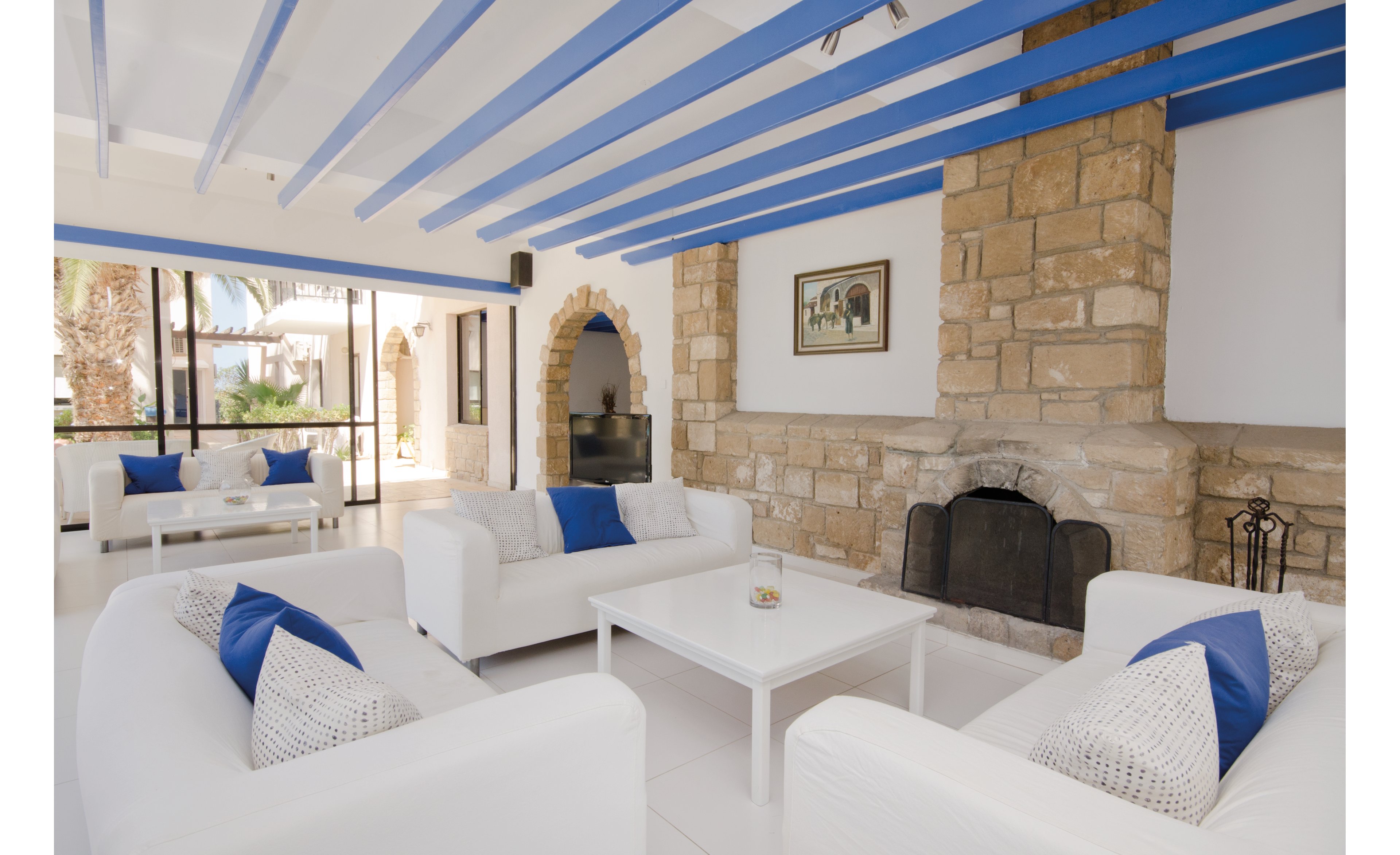 A spacious and airy lounge featuring white furniture with blue cushions, set against a backdrop of stone walls and a fireplace. The room is illuminated by natural light streaming through large windows, showcasing an outdoor view with palm trees. The ceiling beams painted in blue add a vibrant touch to the serene Mediterranean aesthetic.