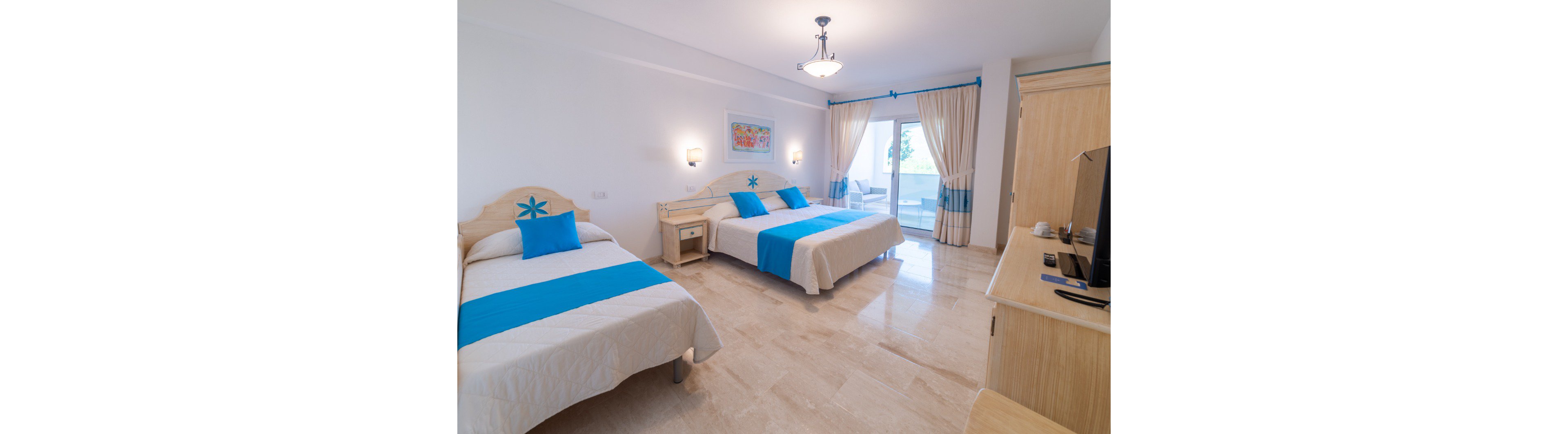 Spacious hotel room featuring a double bed and a single bed with white linens and bright blue accents. The room has light wood furniture, including a bedside table and a cabinet with a television. Large glass doors with cream curtains open to a balcony, allowing natural light to fill the space. Wall-mounted lamps and a ceiling light provide additional illumination, while a colorful artwork decorates the wall.