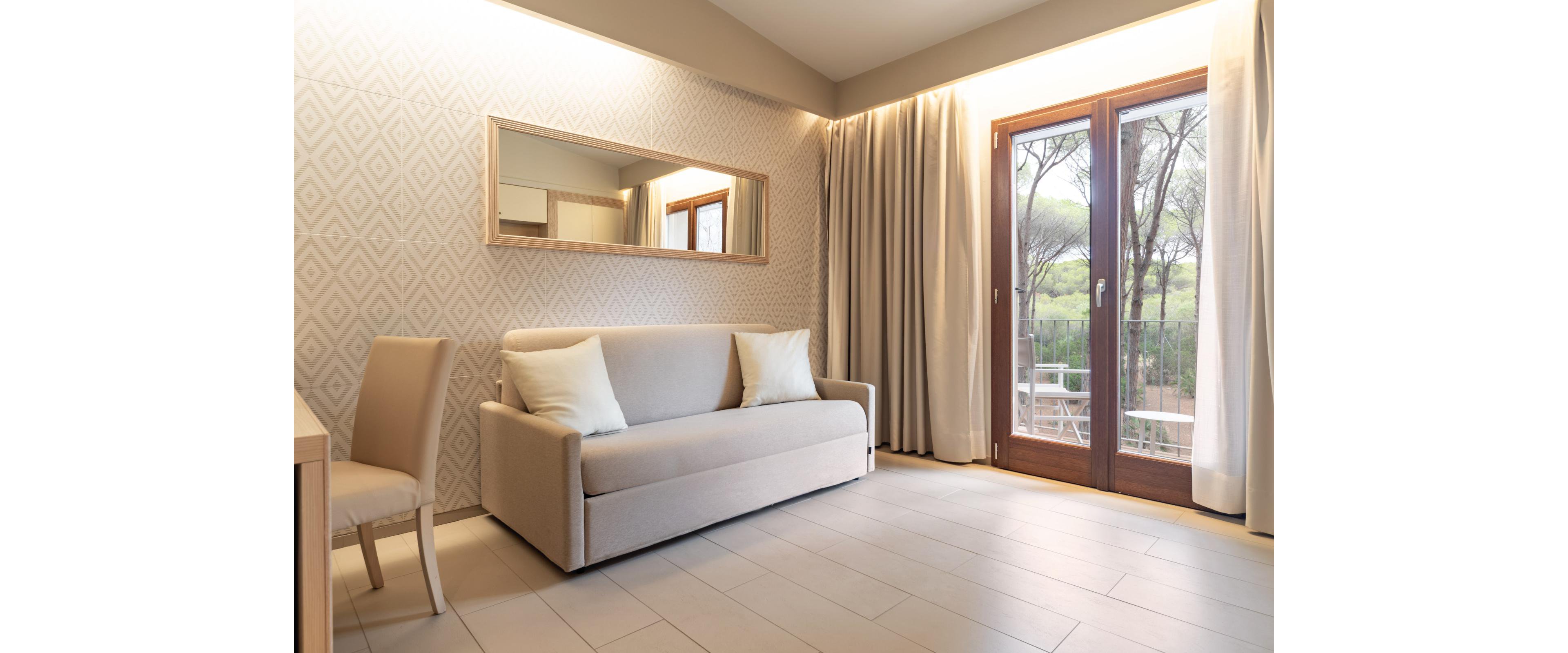 A contemporary hotel room features a beige sofa with white pillows against a patterned accent wall. A simple chair and desk sit to the left, while a large mirror reflects the clean, minimalist decor. Floor-to-ceiling curtains frame glass doors that open onto a balcony with outdoor seating. Trees and greenery are visible outside, adding a calm, natural backdrop to the serene interior.