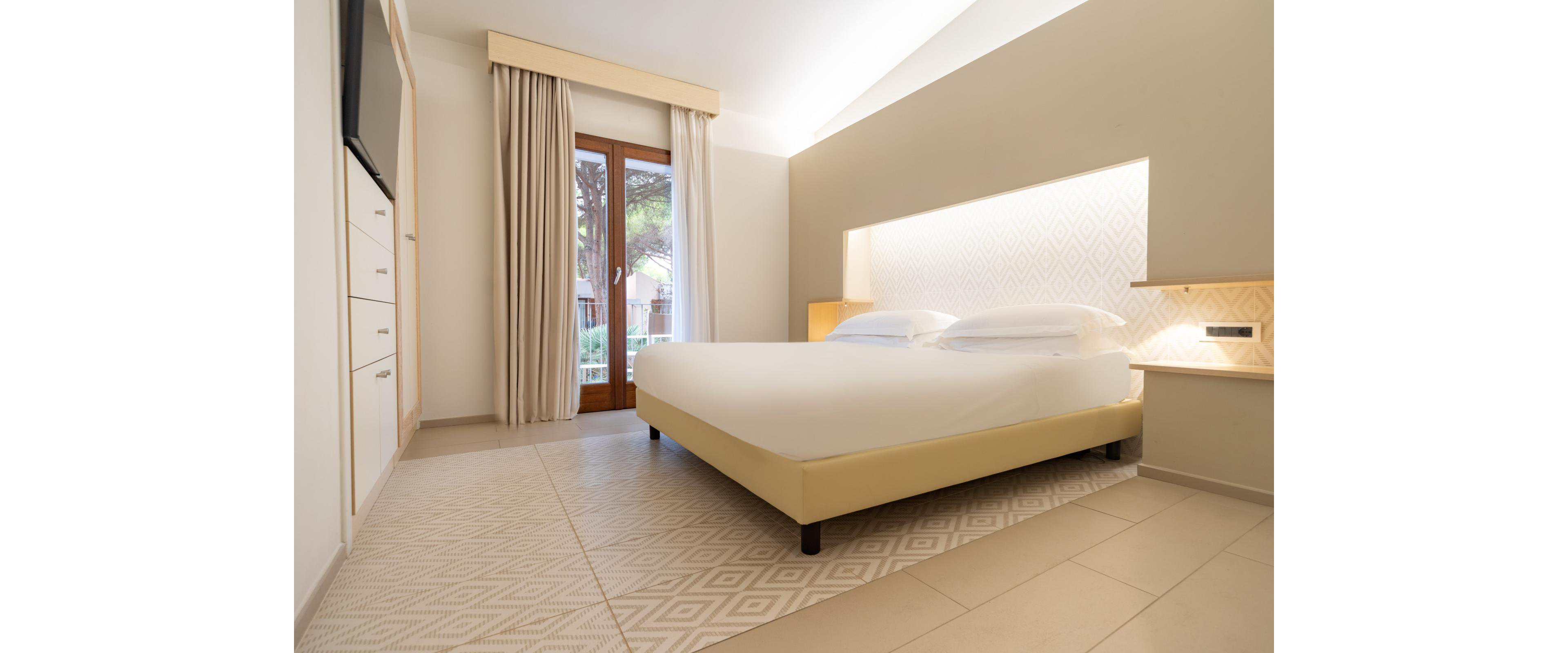 A contemporary hotel bedroom features a large double bed with crisp white linens and a padded beige frame. Soft indirect lighting illuminates the textured headboard wall, creating a warm and inviting atmosphere. Floor-to-ceiling curtains frame a glass door that opens to an outdoor balcony with greenery visible outside. Neutral tiles and minimal furnishings emphasize a clean, spacious, and modern design.