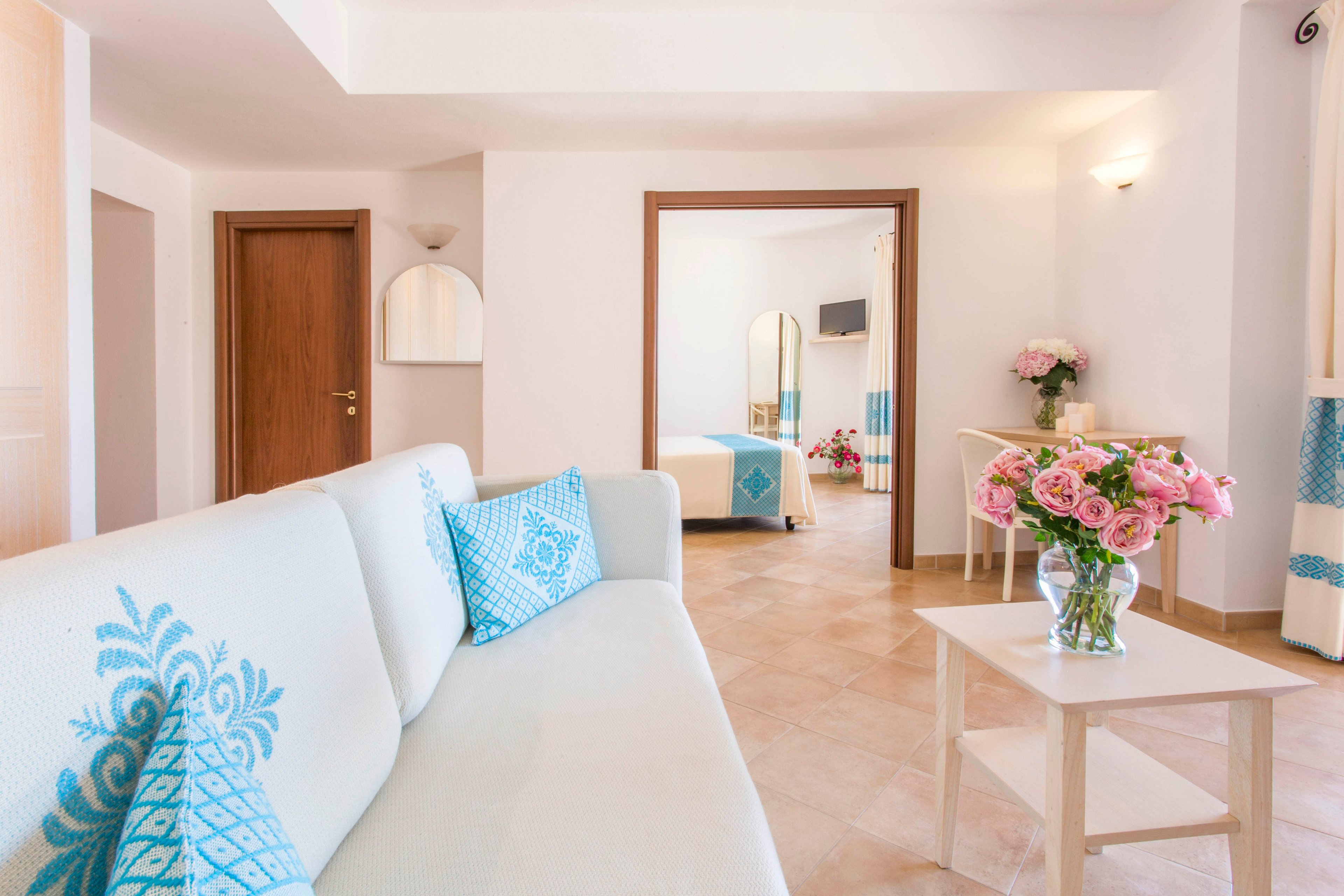 A bright hotel suite features a white sofa with turquoise patterned cushions in the foreground. A small wooden table holds a glass vase filled with pink flowers, adding a soft decorative touch. In the background, an adjoining bedroom with a neatly made bed and matching turquoise accents is visible through an open doorway. Warm tiled floors, white walls, and soft lighting create an inviting, coastal-inspired atmosphere.