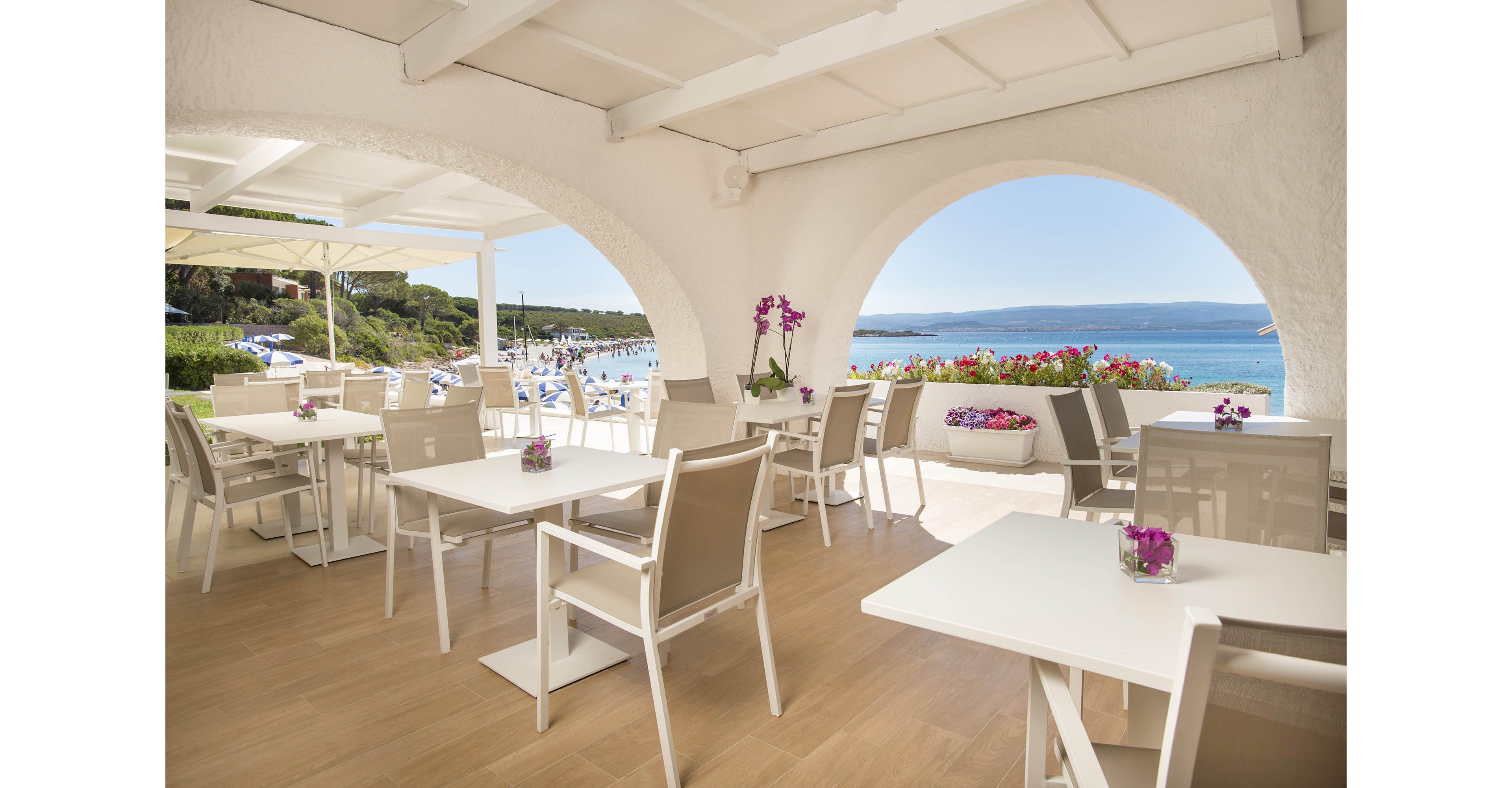 A bright, open-air restaurant terrace overlooks a scenic beach and blue ocean. White tables and chairs are neatly arranged under a shaded pergola, with purple flowers adding a touch of color to the setting. The atmosphere is tranquil and inviting, ideal for relaxed dining by the sea. Large arched openings frame the picturesque coastal landscape. No visible text or numbers are present in the image.