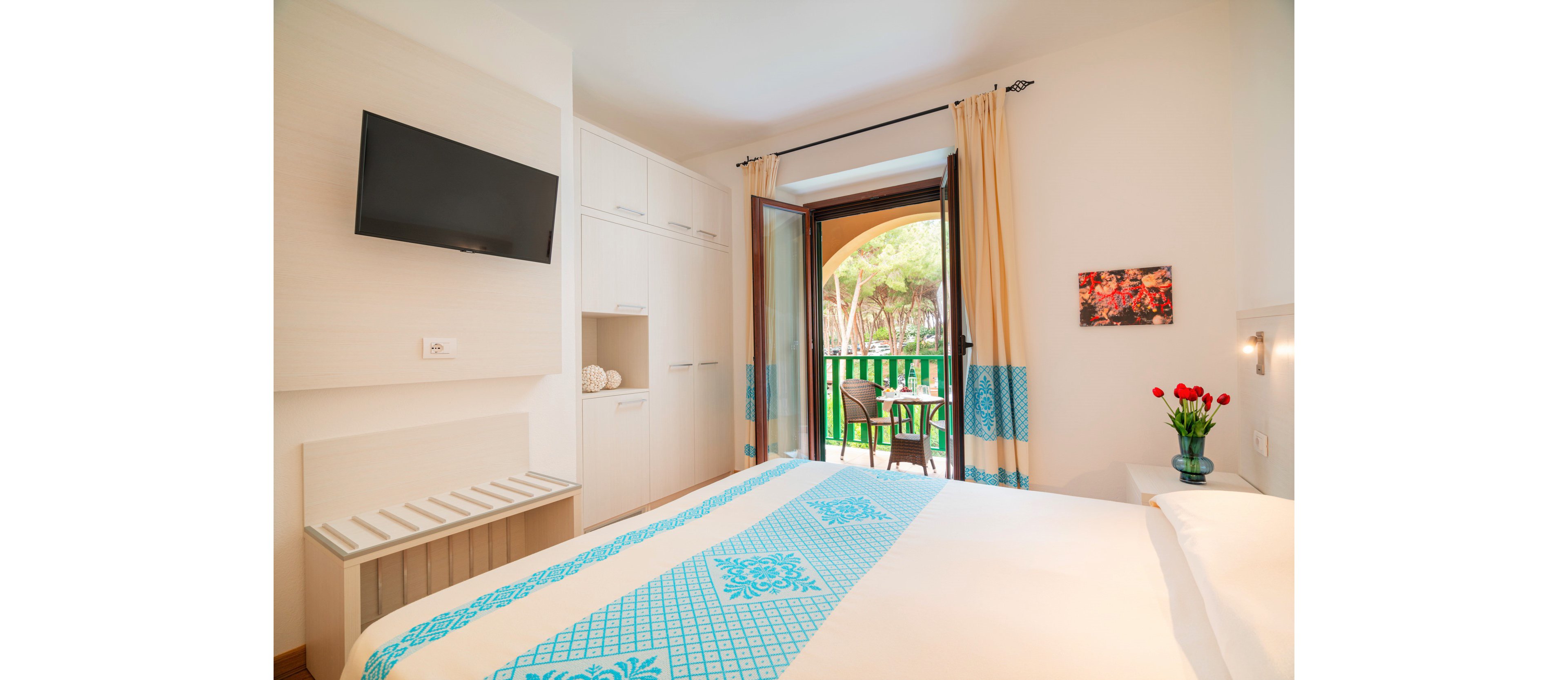 A sunlit hotel room features a large bed with blue patterned linens, a wall-mounted flat screen TV, and built-in white cabinetry. The room opens onto a balcony with outdoor seating, overlooking greenery. Red flowers in a vase and a colorful wall art add vibrant accents to the clean, minimalist space.