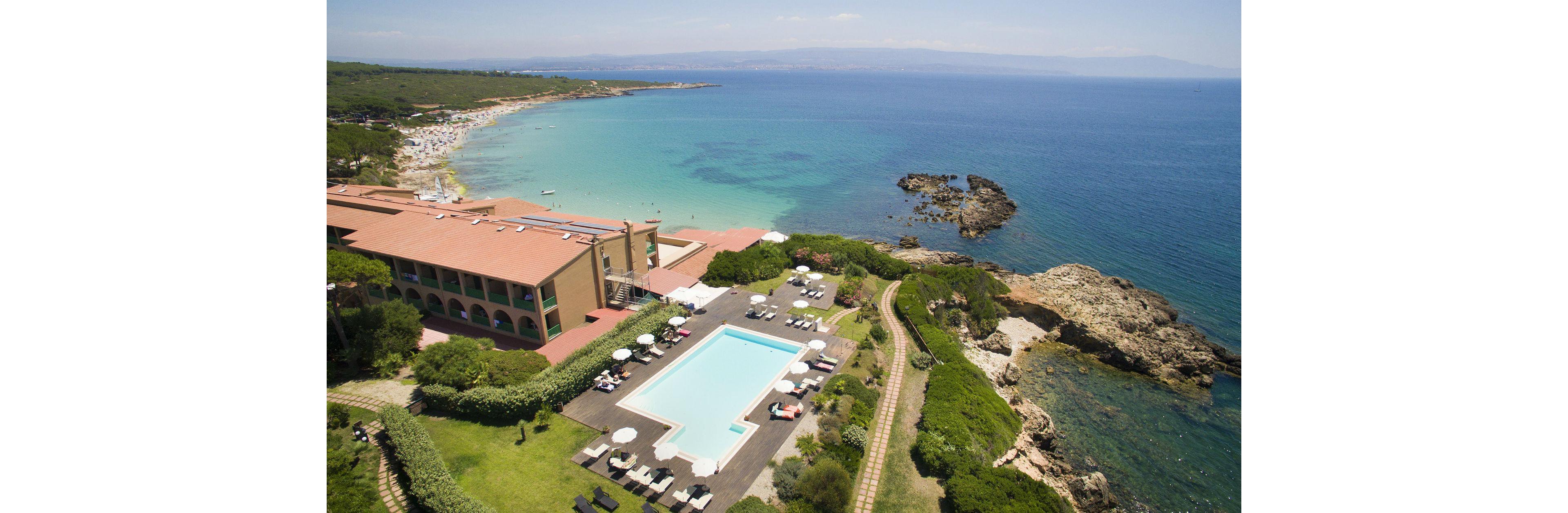 A large seaside resort features a rectangular outdoor swimming pool surrounded by lounge chairs and umbrellas. The property is set against a picturesque coastline with clear blue waters, rocky outcrops, and lush greenery. The beach is visible in the background, with people enjoying the sun and sea. The overall mood is relaxed and inviting, with a bright and natural color palette.