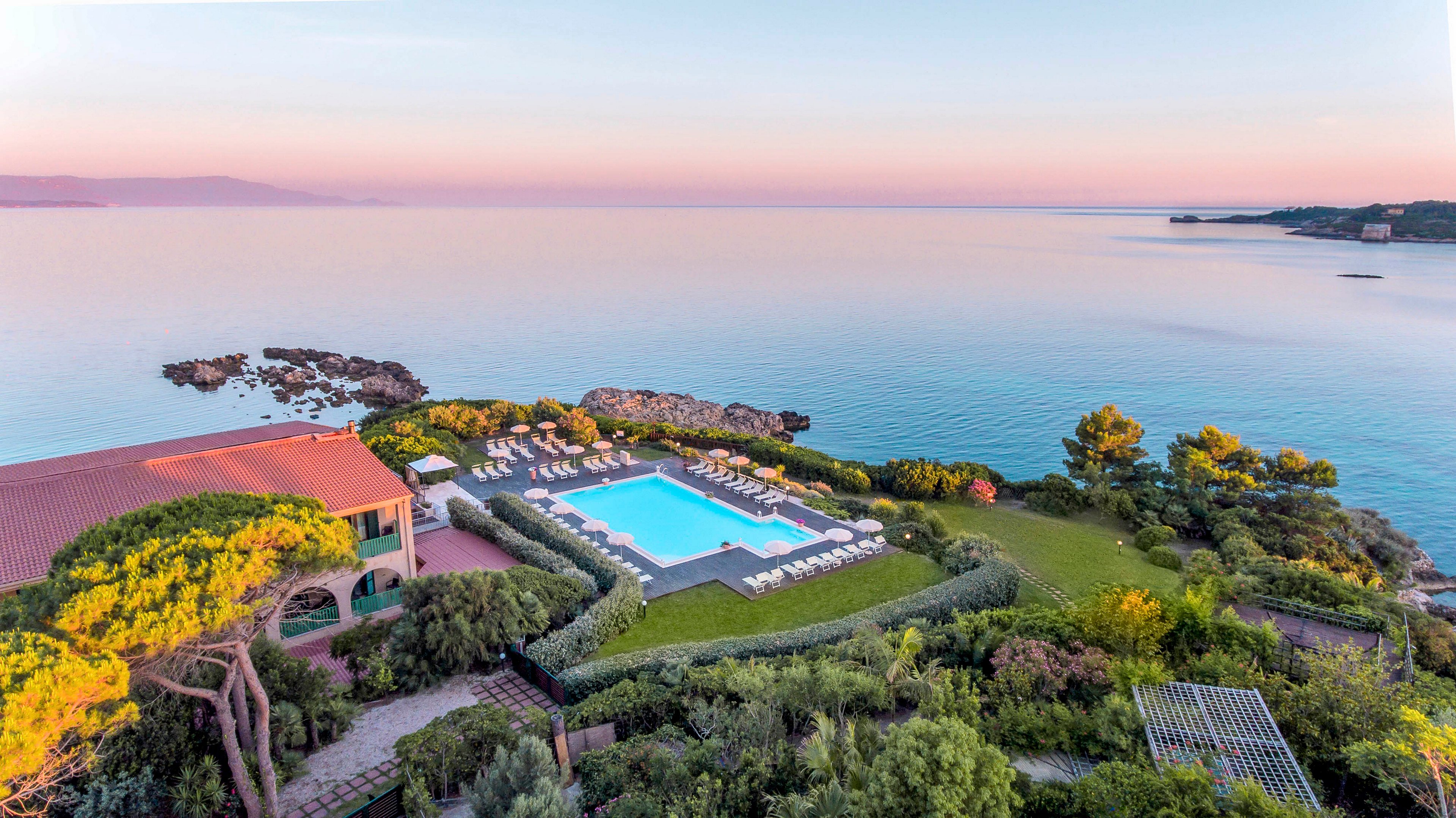 Aerial view of a luxury resort featuring a large rectangular swimming pool surrounded by lounge chairs and lush gardens. The property is set on a scenic coastline with calm, pastel-colored waters and a clear sky at sunrise. The tranquil atmosphere is enhanced by the soft pink and blue hues reflecting off the sea, creating a serene and inviting mood.