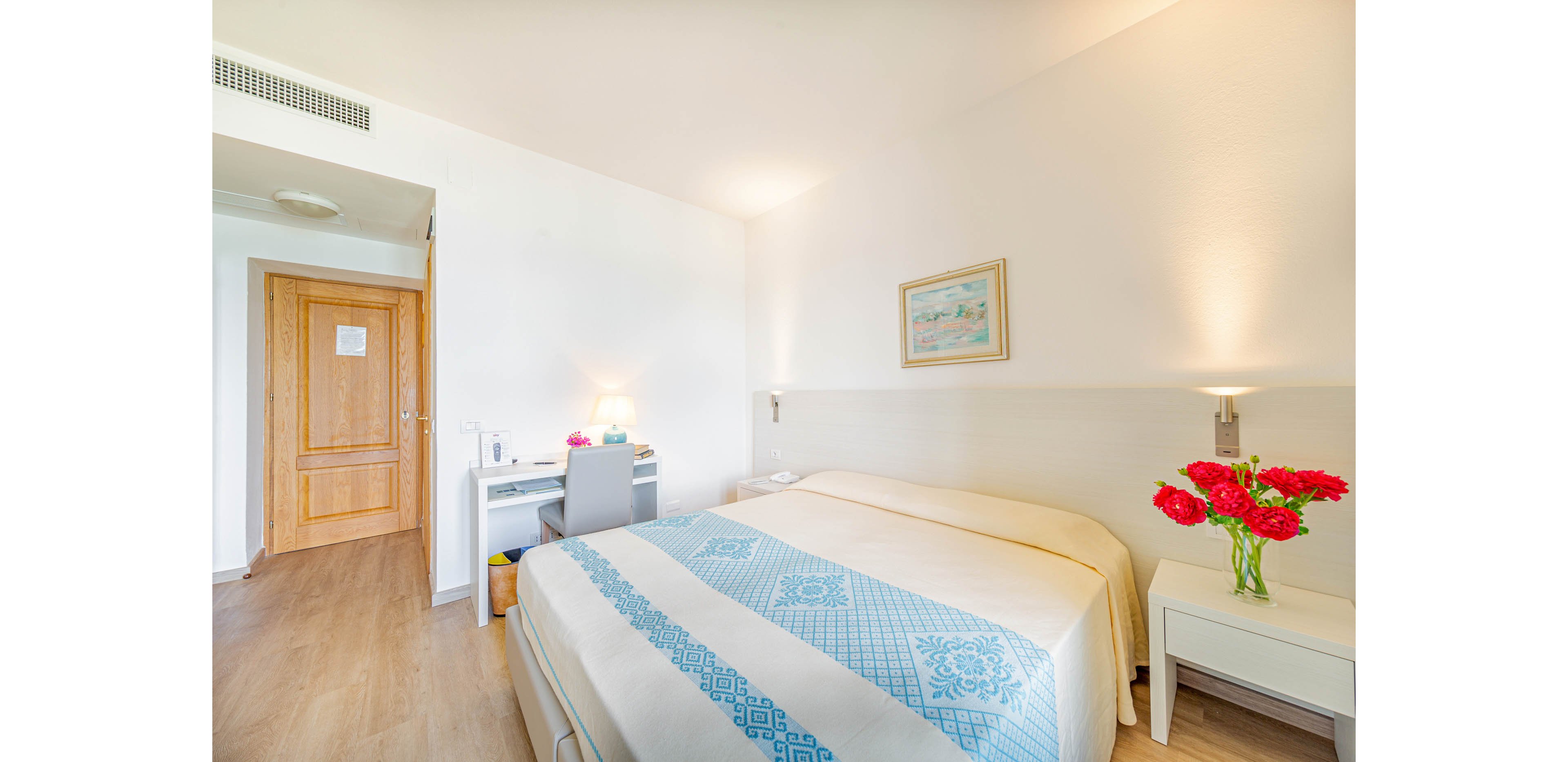 A spacious hotel room features a large double bed with a blue and white patterned blanket. The setting includes a wooden floor, a desk with a chair and lamp, and a nightstand with a vase of red flowers. Soft lighting and minimal decor create a calm, inviting atmosphere. A framed artwork hangs above the bed, and a door with a visible sign leads to the hallway.