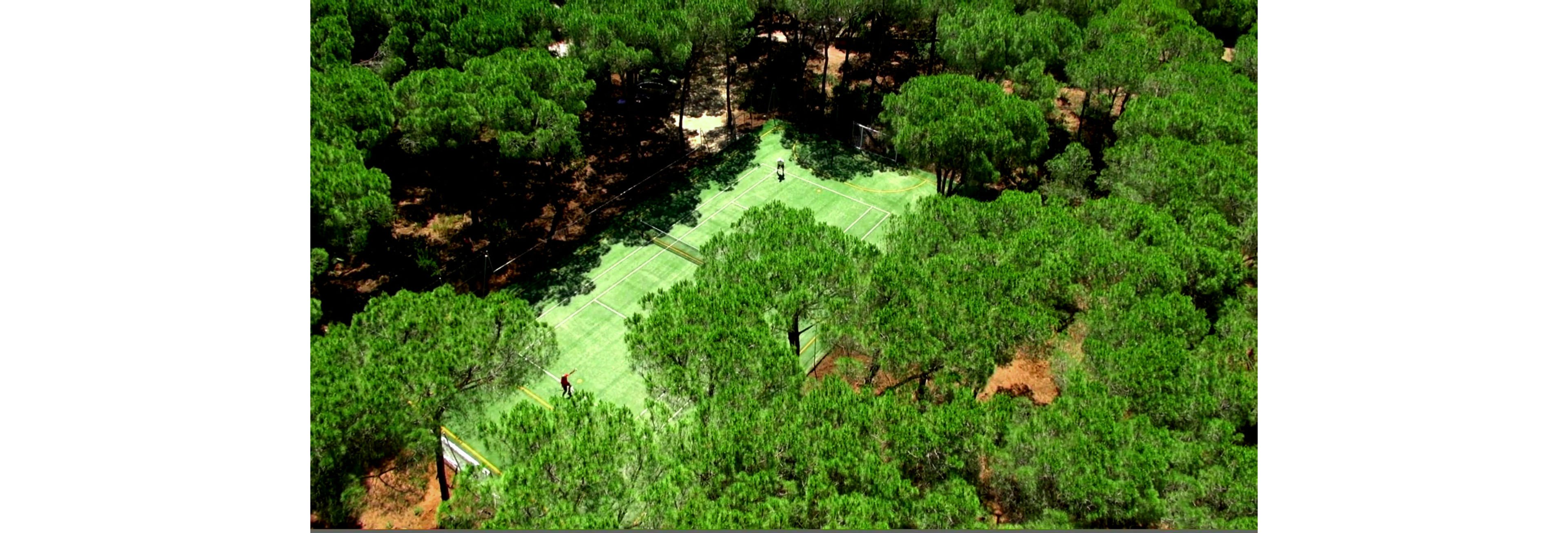 This image shows a tennis court nestled within a dense forest, viewed from above. Two people are visible on the court, engaged in a game or practice session. The lush green trees dominate the scene, creating a striking contrast with the lighter green surface of the court. The setting appears tranquil and secluded, emphasizing nature and outdoor recreation.
