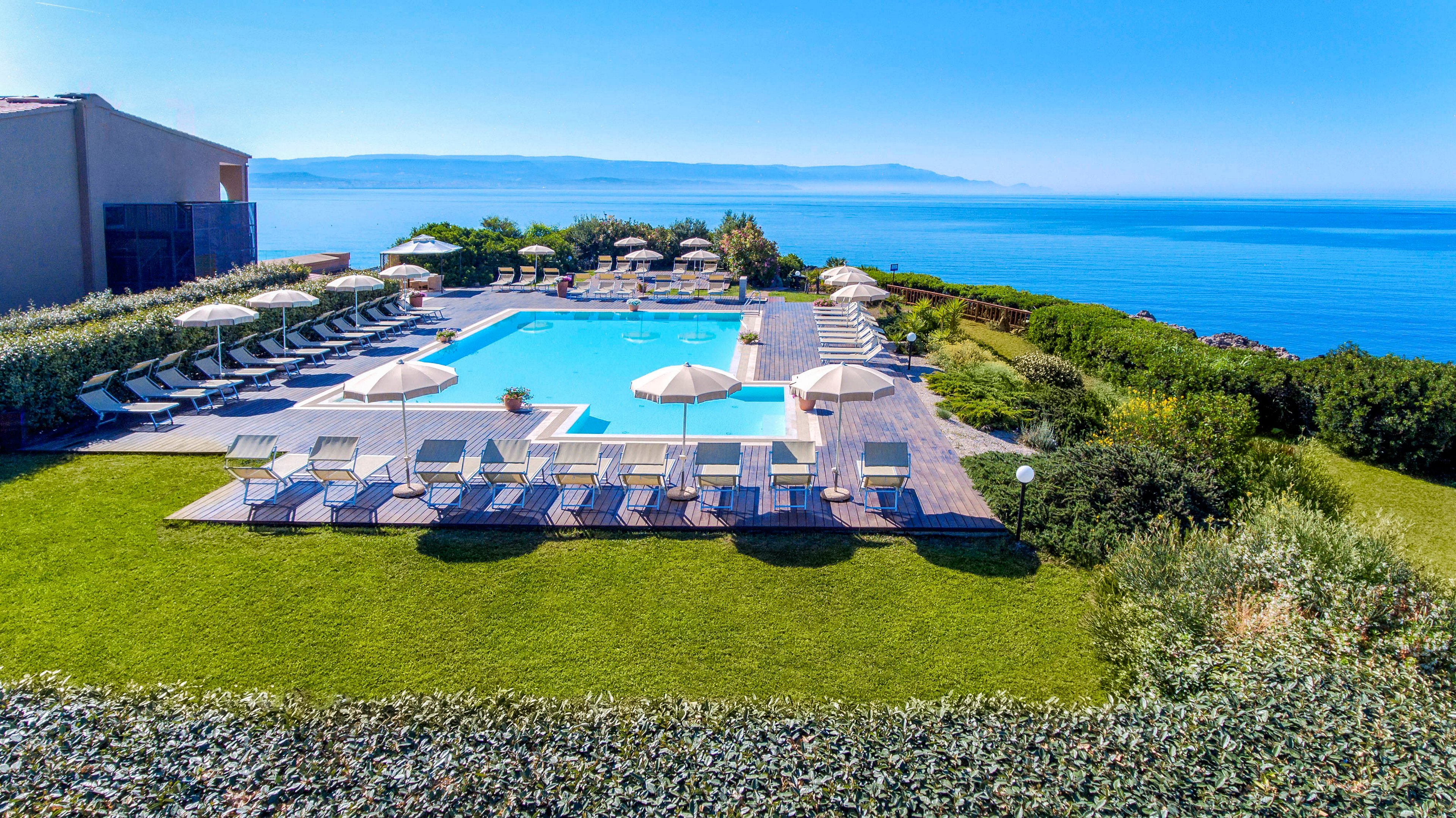 A modern outdoor swimming pool is surrounded by numerous sun loungers and umbrellas, set on a manicured lawn overlooking a stunning blue sea. The setting is tranquil and inviting, with clear skies and lush greenery enhancing the relaxing atmosphere. The pool area is elevated, providing panoramic ocean views and a serene vacation vibe.