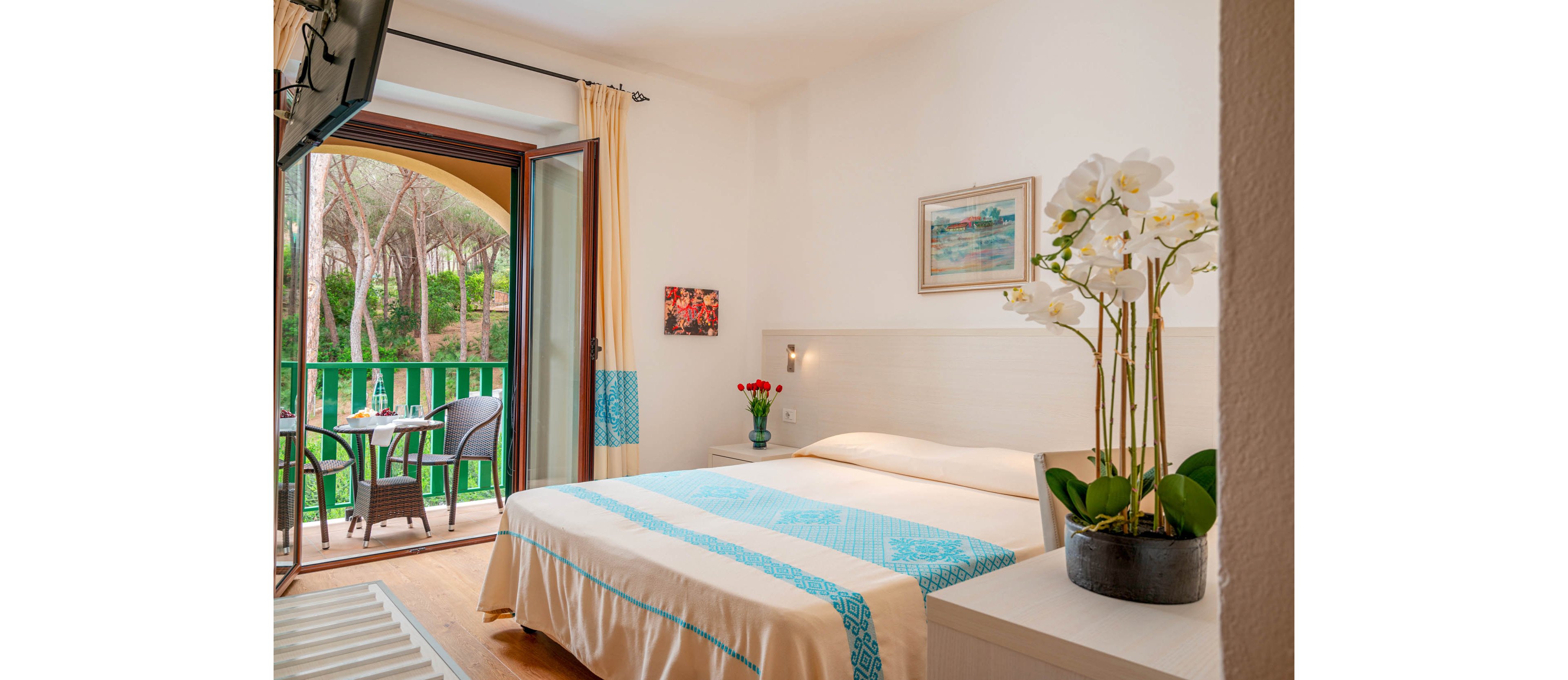 A sunlit hotel room features a neatly made double bed with turquoise accents and a large potted orchid on the bedside table. The room opens onto a balcony with two chairs and a table, overlooking a lush green garden with tall trees. The decor is modern and minimal, with light walls, framed artwork, and fresh flowers adding a welcoming touch.