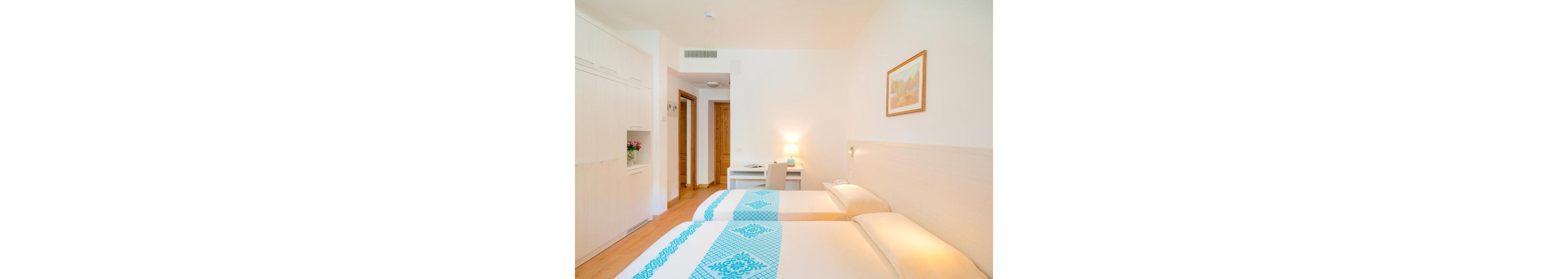 A clean, modern hotel room features two neatly made twin beds with white linens and blue patterned runners. The room includes a small desk with a lamp, a chair, and a vase of flowers, creating a welcoming workspace. Warm wooden doors and minimalistic decor contribute to a calm, inviting atmosphere. A framed artwork hangs on the wall, and soft lighting enhances the serene mood.