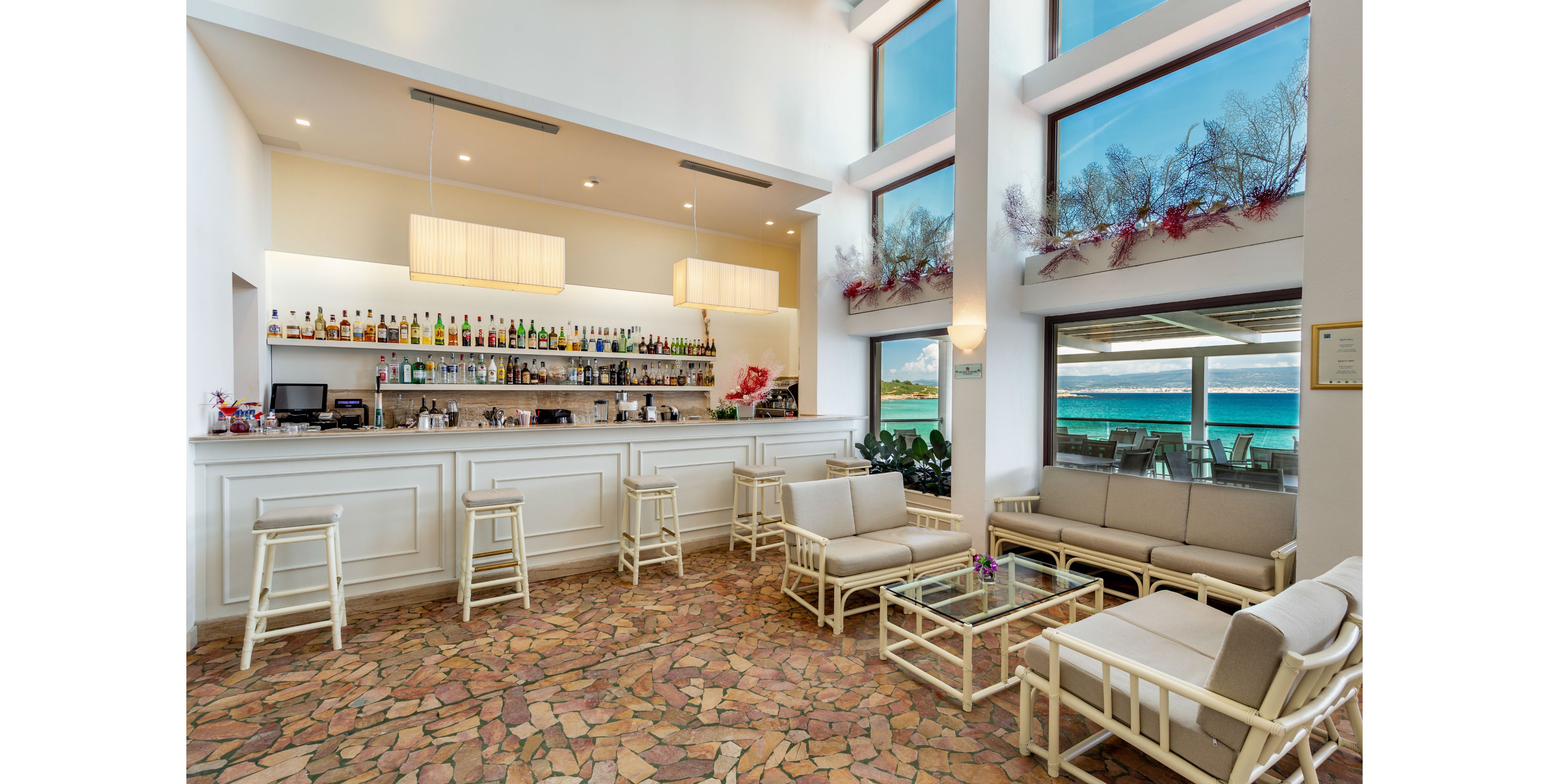 A contemporary bar lounge features white furniture and a well-stocked bar set against large windows overlooking a turquoise ocean. The seating area includes cushioned chairs and glass tables, with natural light flooding the space. Decorative plants and festive garlands add a touch of color, while the stone-tiled floor enhances the relaxed, coastal atmosphere.