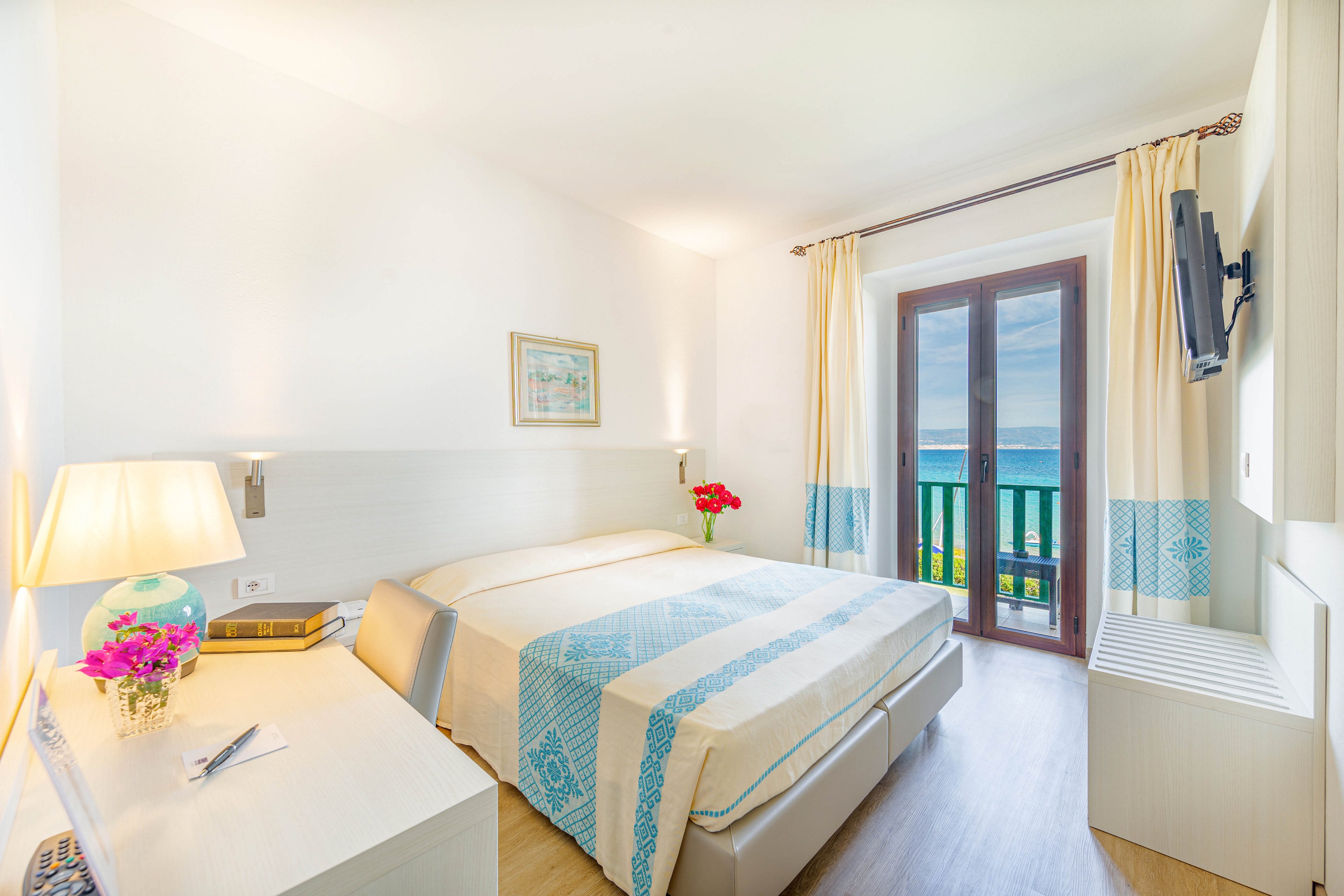 A sunlit hotel room features a double bed with blue and beige linens, a writing desk, and a wall-mounted TV. Large glass doors open to a balcony overlooking the ocean, bringing in natural light. The decor is minimal and fresh, with flowers and a lamp adding warmth to the modern space.