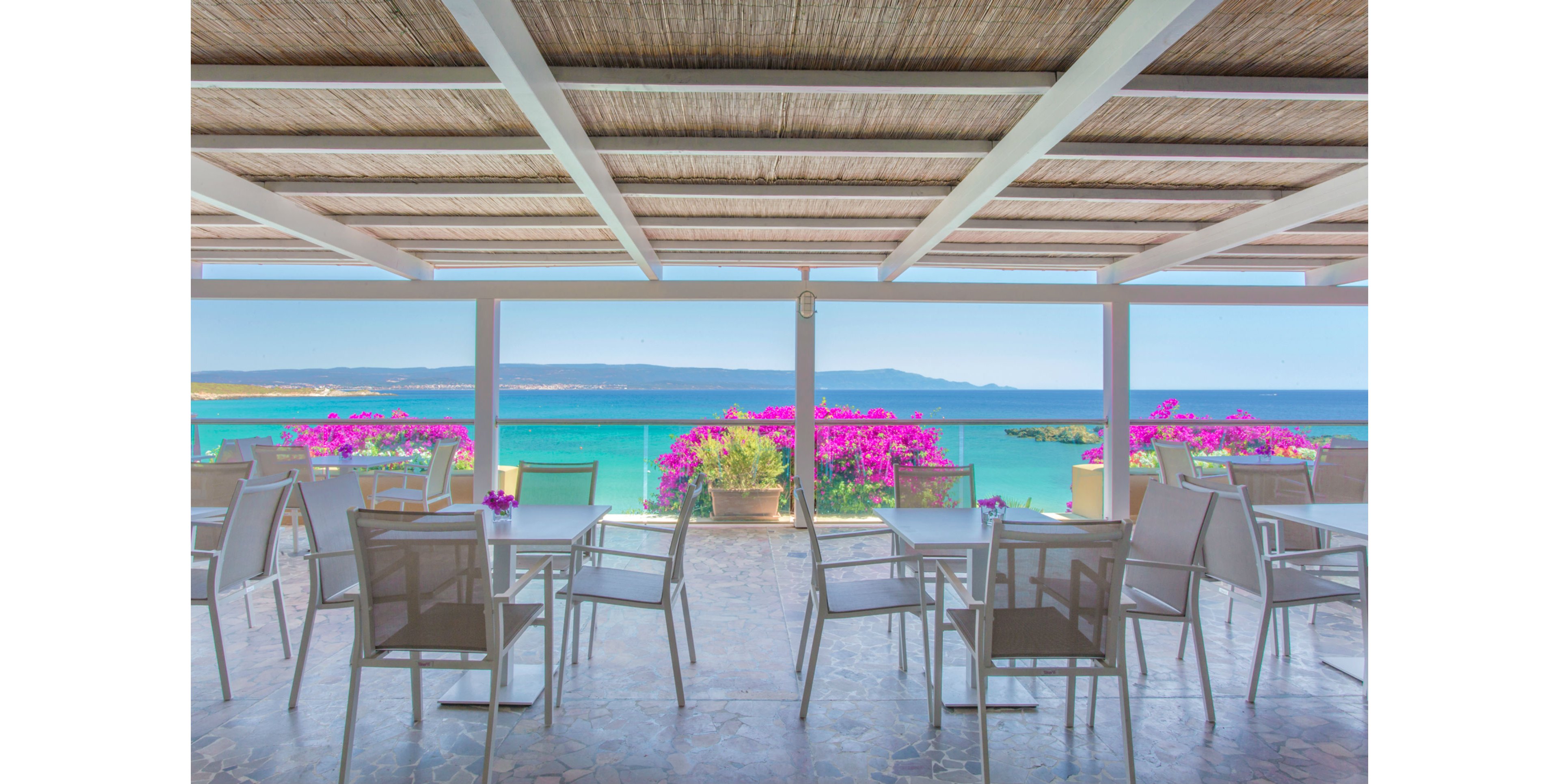 An open-air terrace café overlooks a turquoise ocean, framed by vibrant pink bougainvillea flowers in planters. The seating area features modern white tables and chairs under a wooden pergola roof, creating a relaxed and inviting atmosphere. The clear blue sky and distant mountains enhance the tranquil coastal setting. No people are present, emphasizing the peaceful mood. No visible text or numbers appear in the image.