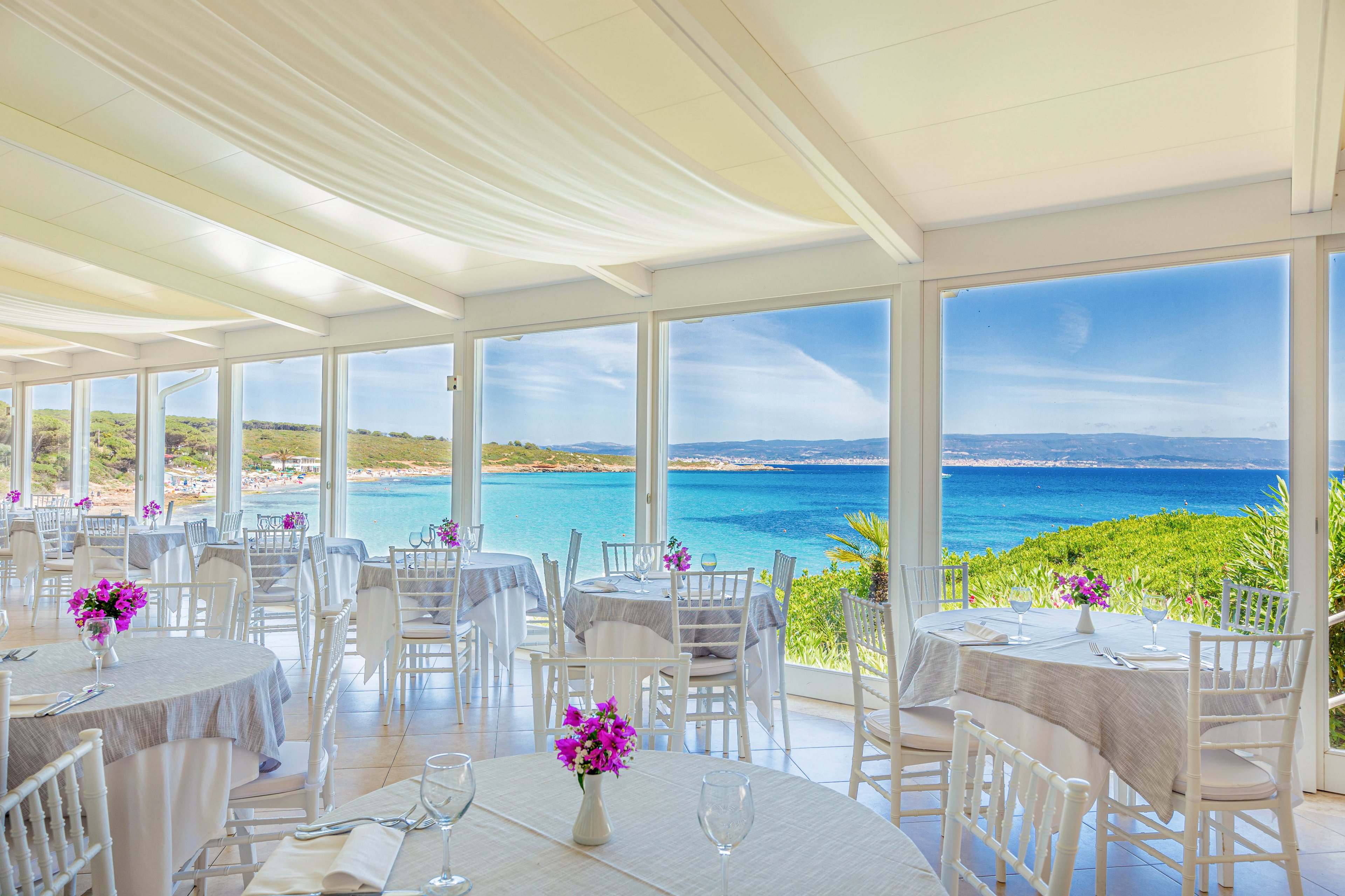 The image features a bright, airy event space with large windows overlooking a stunning blue ocean and coastline. Round tables are set with white linens, glassware, and vibrant purple floral centerpieces, creating a festive and elegant atmosphere. The setting suggests a daytime celebration or gathering, with natural light flooding the room and a serene, coastal landscape visible outside. The mood is cheerful and inviting, enhanced by the fresh color palette and scenic backdrop.