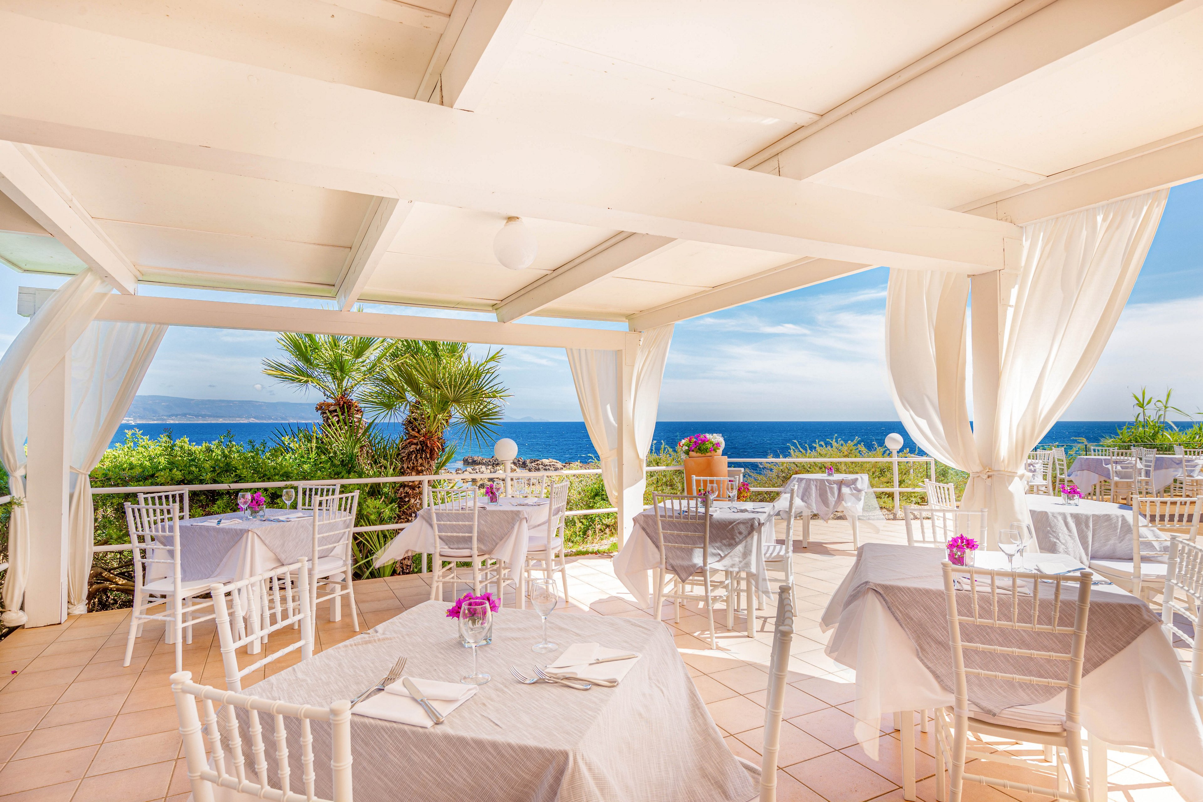 A bright, open-air restaurant terrace overlooks a stunning blue ocean, with white tables and chairs arranged neatly under a shaded pergola. The setting features flowing white curtains, potted plants, and small purple floral centerpieces on each table. The mood is serene and inviting, with natural sunlight and a soft pastel color palette enhancing the relaxing coastal ambiance.