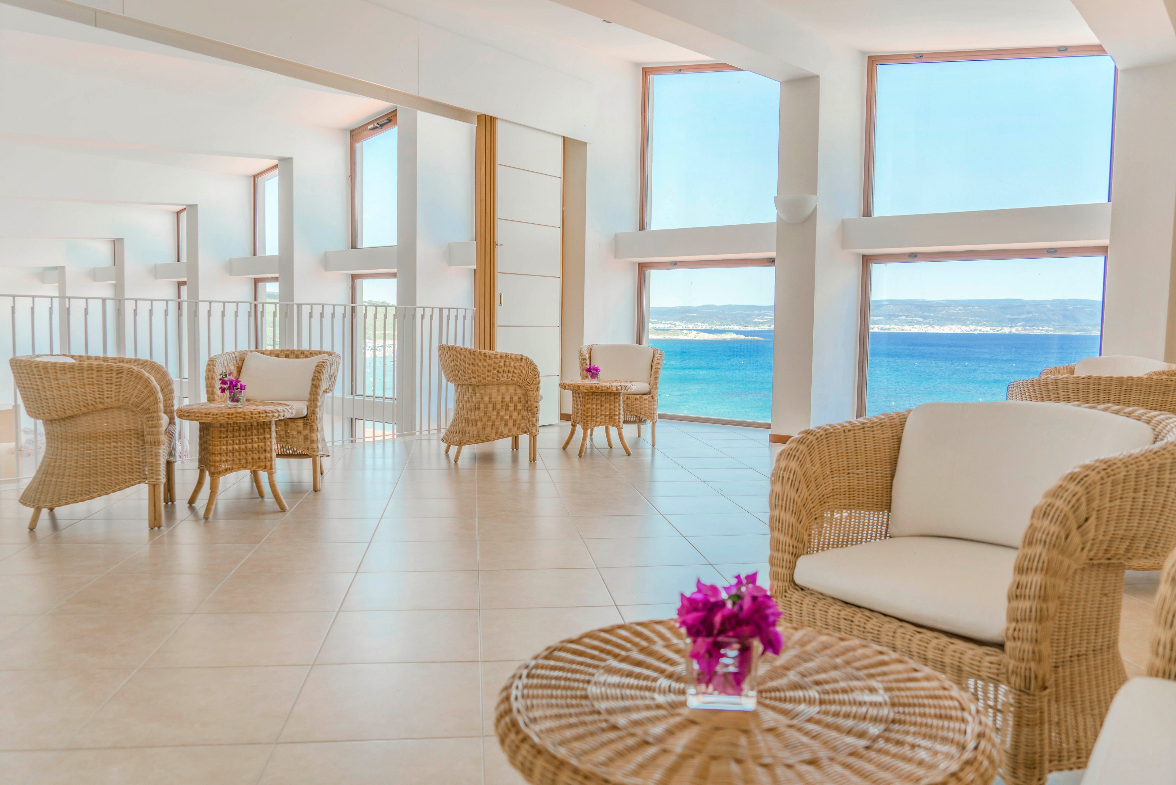 The image features a spacious lounge area with wicker chairs and tables, each adorned with a small purple floral arrangement. Large windows offer a stunning view of the blue ocean and sky, filling the room with natural light. The setting is modern and airy, with a neutral palette and minimalist decor, creating a relaxed and inviting atmosphere. No visible text or numeric values are present.