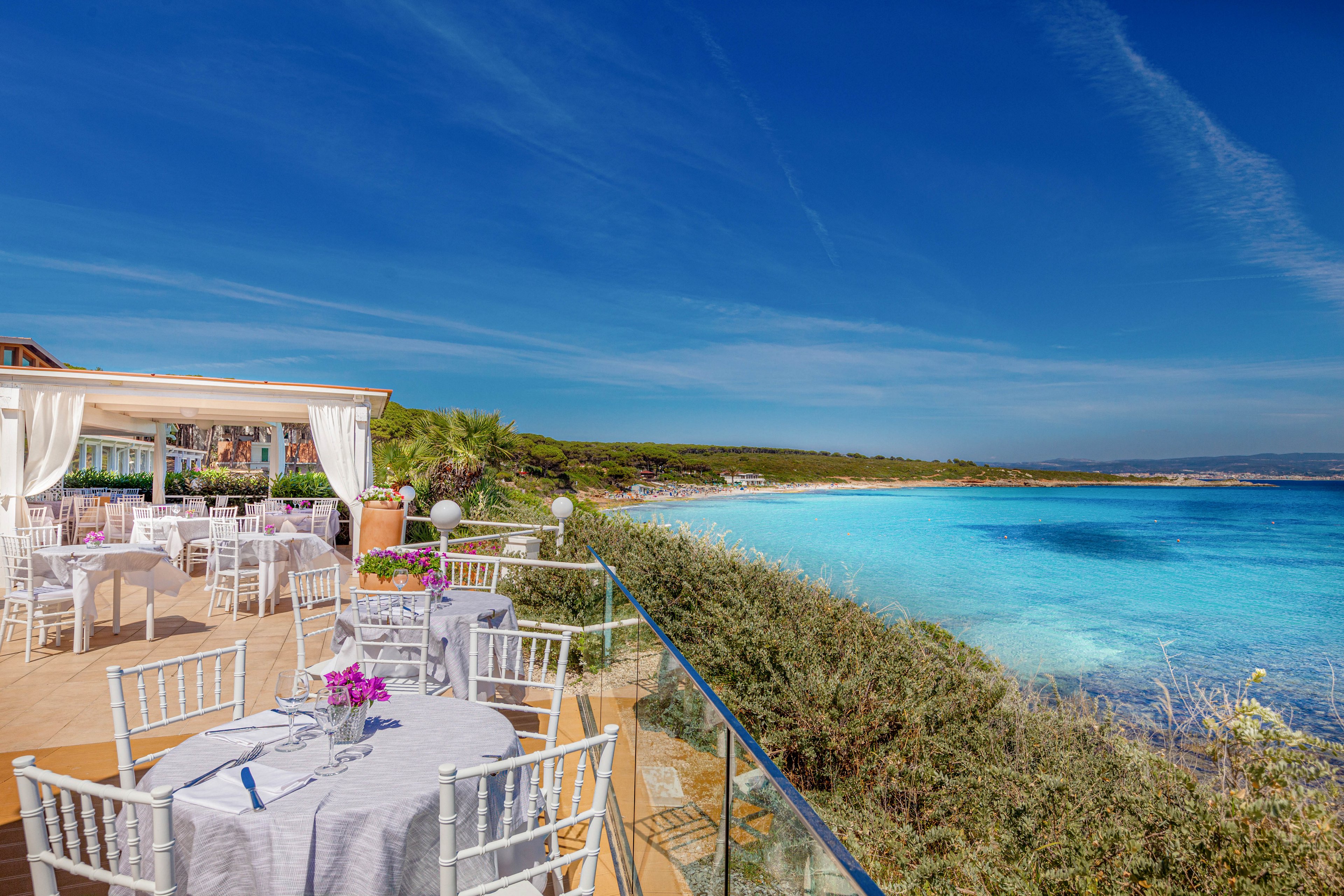 The main subject is a beautifully arranged outdoor dining area with white chairs and tables, some adorned with purple flowers. The setting is a coastal terrace with clear blue skies and vibrant turquoise sea, creating a serene and luxurious atmosphere. The visuals highlight the contrast between the crisp white furniture and the lush greenery, with the expansive beach and water providing a stunning backdrop. No visible text or numeric values are present in the image.