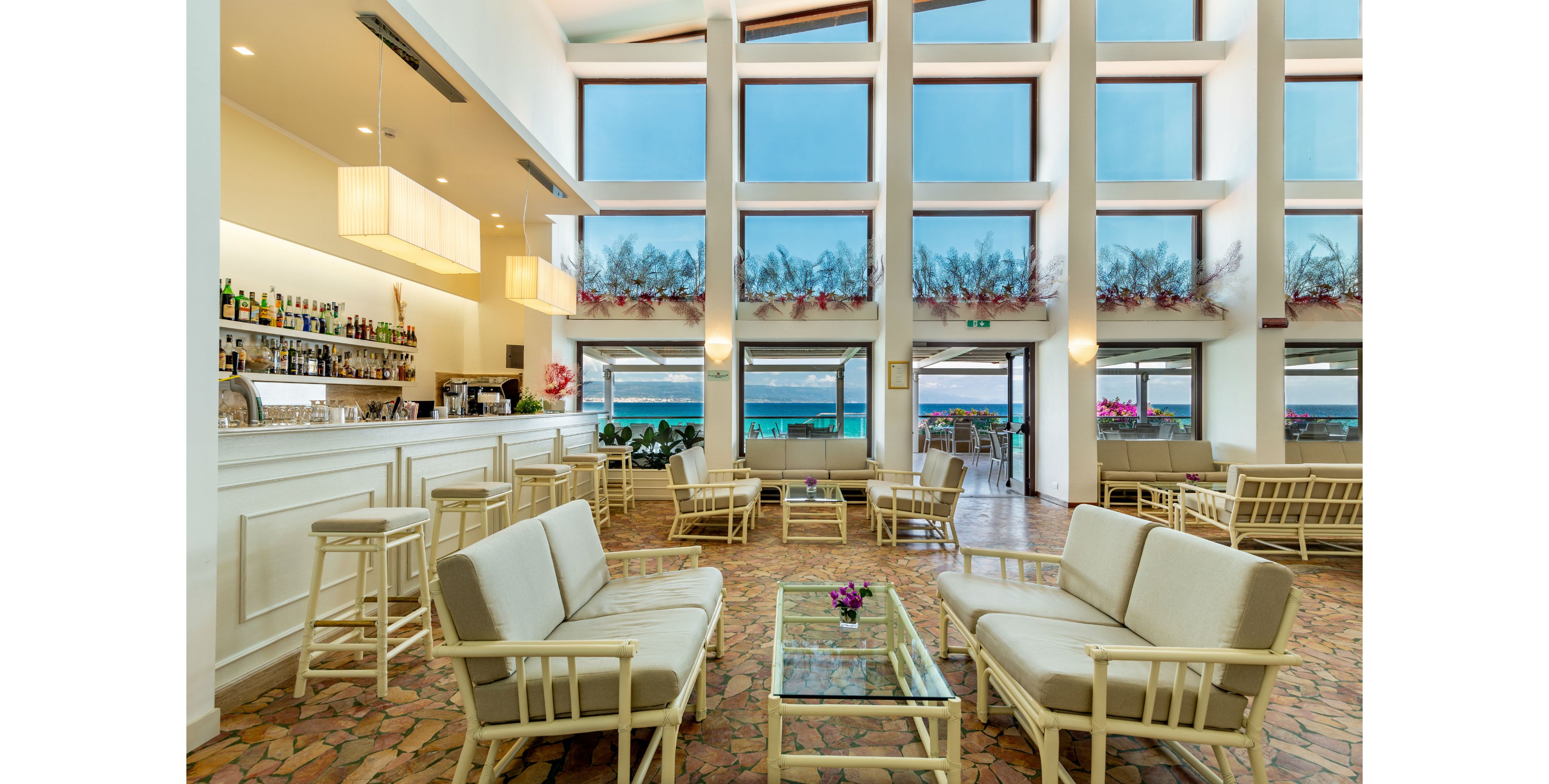 The image features a spacious lounge area with large floor-to-ceiling windows overlooking a scenic beach. The setting includes a white bar with various bottles, cushioned seating, glass tables, and decorative floral arrangements. Natural light fills the room, highlighting the airy and relaxed atmosphere. No visible text or numeric values are present in the image.