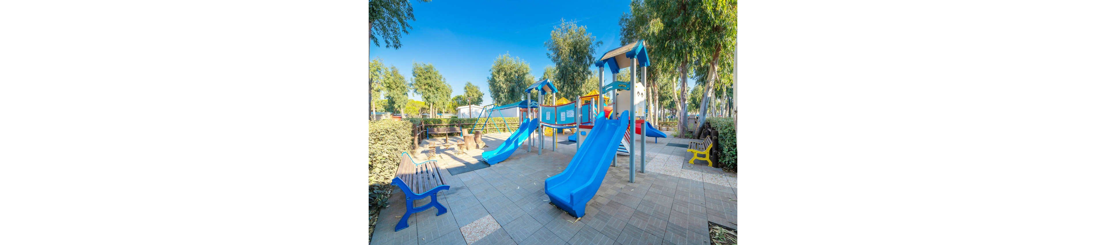 A bright outdoor playground features multiple blue plastic slides and climbing structures. The play area is set in a park surrounded by trees, shrubs, and paved walkways under a clear blue sky. Benches and a small yellow seat provide resting spots around the equipment. The scene appears calm and empty, emphasizing the colorful structures and clean, well-maintained environment.