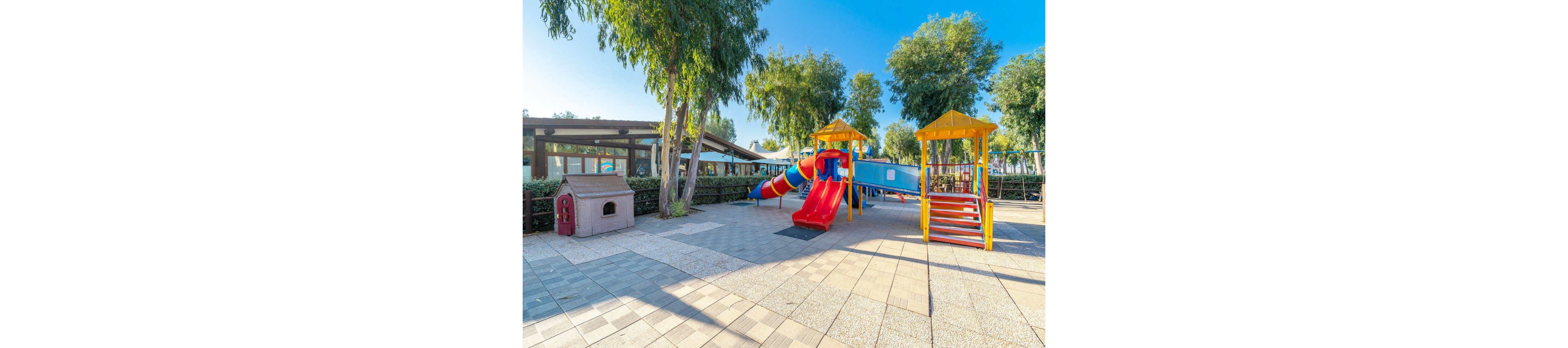 An empty outdoor playground features bright red and yellow slides and climbing structures under a clear blue sky. The play area is surrounded by tall green trees and bordered by a fence. A small playhouse stands to the side on a paved surface with patterned tiles. In the background, a building with a shaded patio suggests a park or recreational facility.