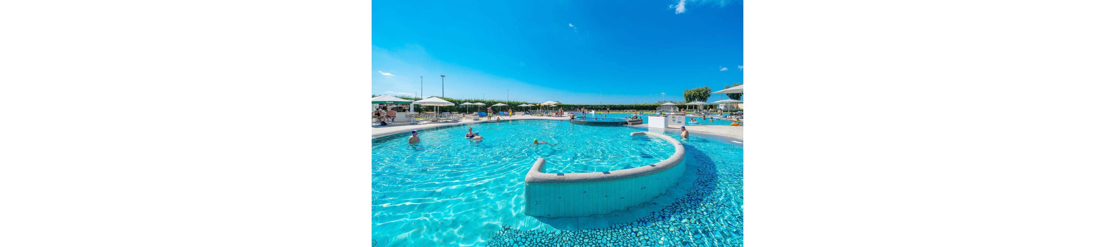 An expansive outdoor swimming pool sparkles under a bright blue sky at a resort. Adults and children relax and play in the clear water while others lounge on deck chairs under white umbrellas. The pool features curved edges, a shallow ledge, and a central circular island area. Green hedges and distant structures frame the leisure scene, creating a relaxed vacation atmosphere.