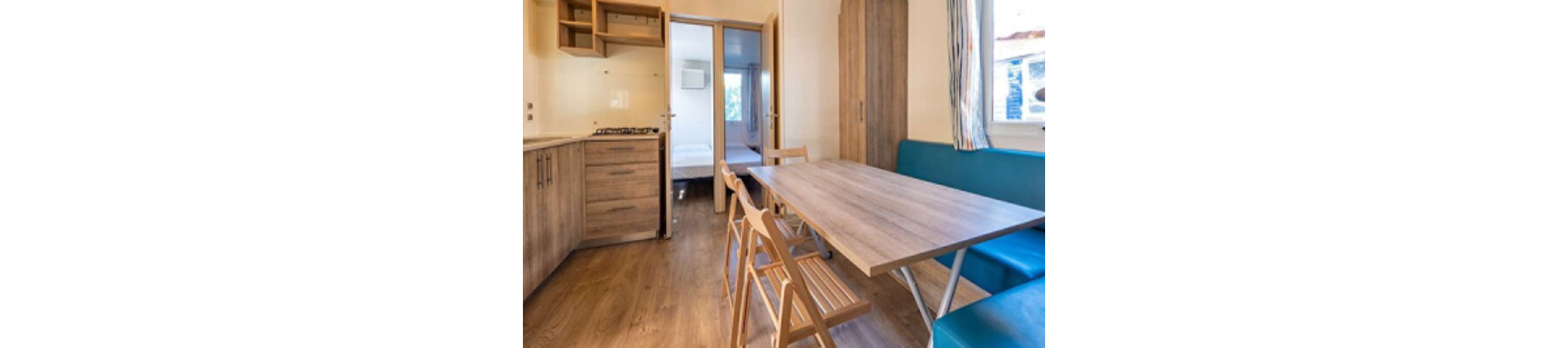 Interior view of a compact holiday cabin featuring a small kitchen and dining space. A wooden table with folding chairs and a turquoise bench sits in the center of the room. Built-in wooden cabinets, a countertop, and a gas cooktop line one wall. Open doors reveal a bedroom in the background, while a window brings in natural light and shows a glimpse of the outdoors.