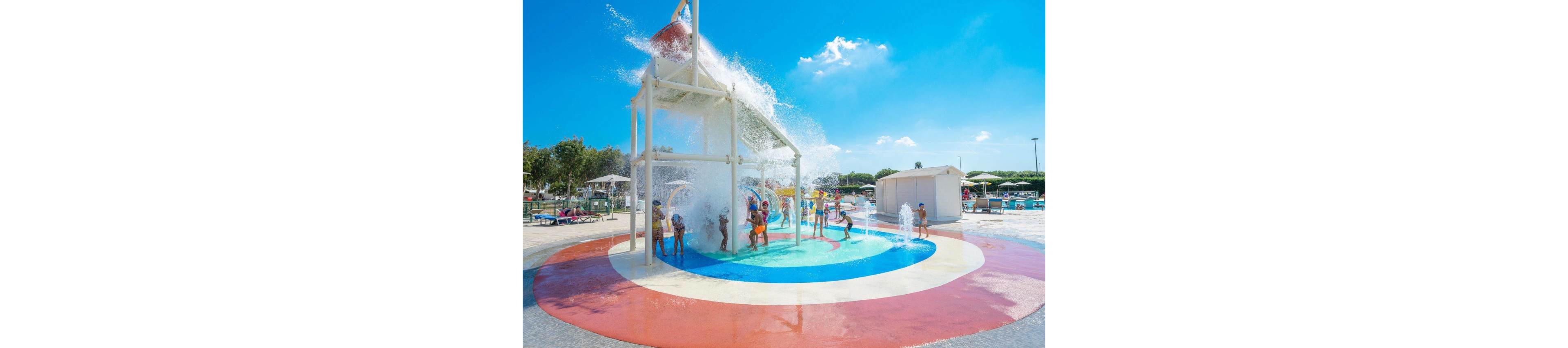 Children play in a colorful splash pad under a large tipping water bucket structure. Water cascades down as kids run through fountains and sprays on the wet surface. The scene is set in an outdoor water park with lounge chairs, umbrellas, and trees in the background. Bright sunlight and a clear blue sky create a vibrant, summery atmosphere.