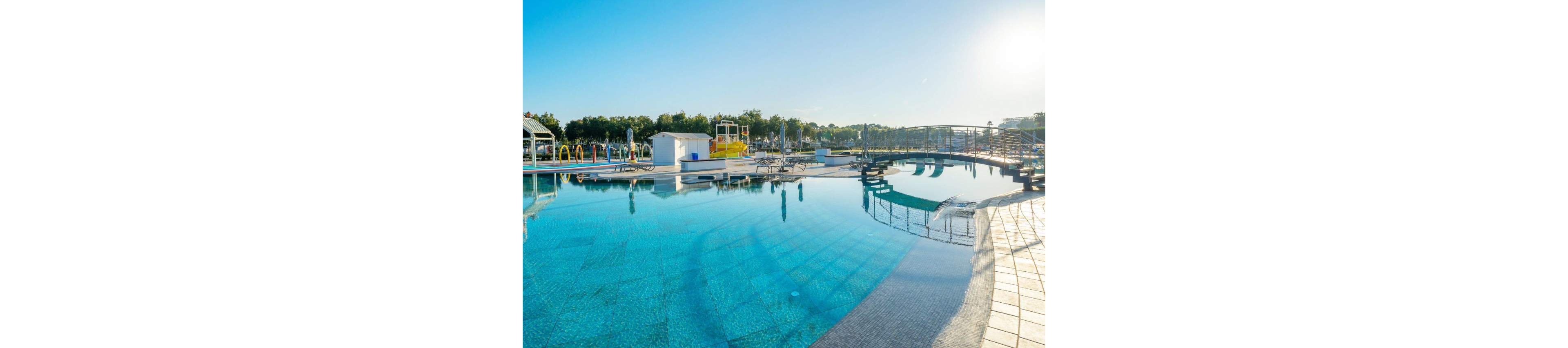 A large outdoor swimming pool reflects the clear blue sky in bright morning sunlight. The scene includes poolside loungers, umbrellas, and small structures around the water. A curved bridge and tiled deck add visual interest and lead the eye across the pool. Trees and greenery in the background suggest a relaxing resort or recreational setting.