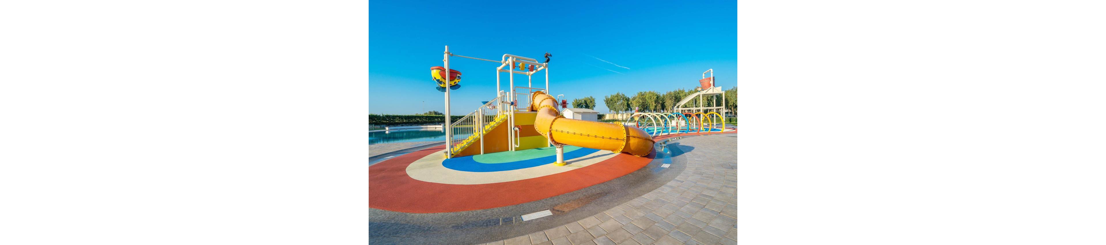 A bright yellow tube slide and splash structure stand in an outdoor water playground. The play area features colorful arches, stairs, and water sprayers on a smooth, wet surface. A calm body of water and green trees are visible in the background under a clear blue sky. The scene suggests a sunny, family-friendly recreational space designed for children.