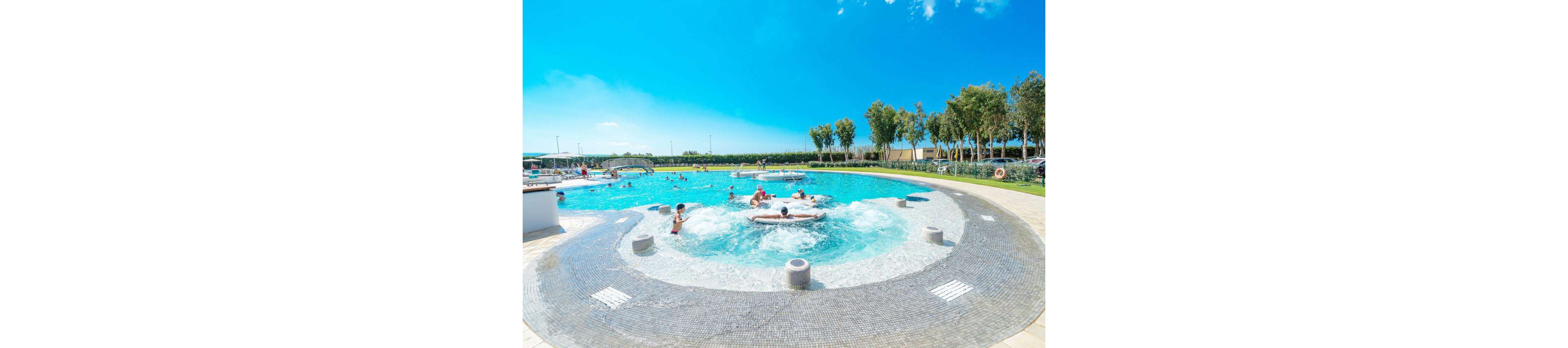 An outdoor resort-style swimming pool features a circular whirlpool area in the foreground with people relaxing in the bubbling water. Swimmers and families enjoy the larger pool behind it under a bright blue sky. Trees and landscaped greenery line the edge of the pool area, creating a vacation atmosphere. The scene suggests leisure, summer fun, and a family-friendly water park environment.