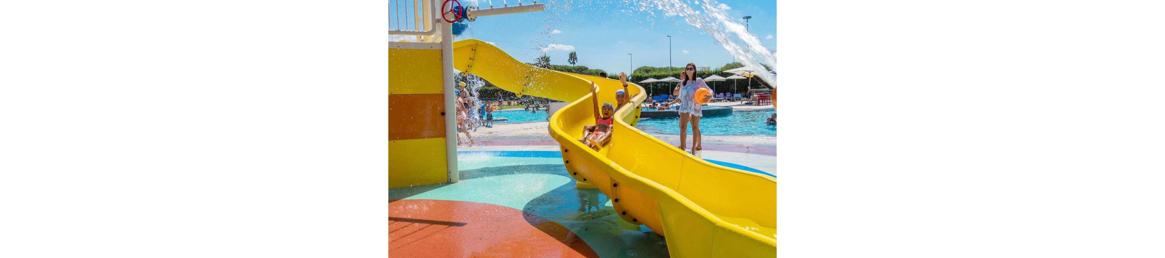 Two children ride down a bright yellow waterslide at an outdoor water park. Water splashes around them as they curve toward a shallow pool. In the background, adults and other visitors relax near umbrellas and lounge chairs. The scene is sunny and colorful, emphasizing summer fun and family recreation.