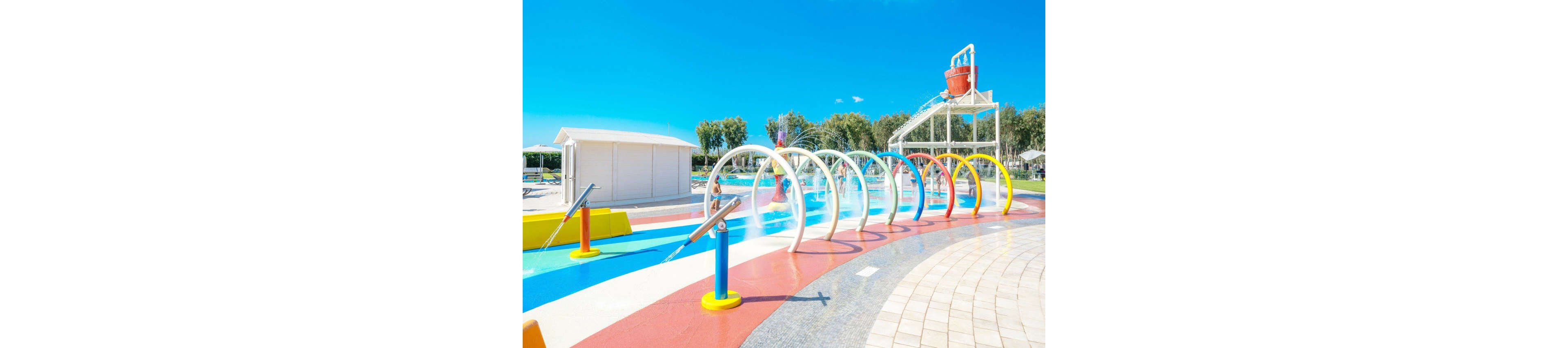 A bright outdoor splash park features multiple colorful water arches spraying over a shallow play area. The ground is painted in bold red, blue, and white stripes leading toward a larger water structure. A tipping bucket and additional spray elements stand in the background under a clear blue sky. Trees and a small white building frame the aquatic playground, creating a fun and inviting summer setting.