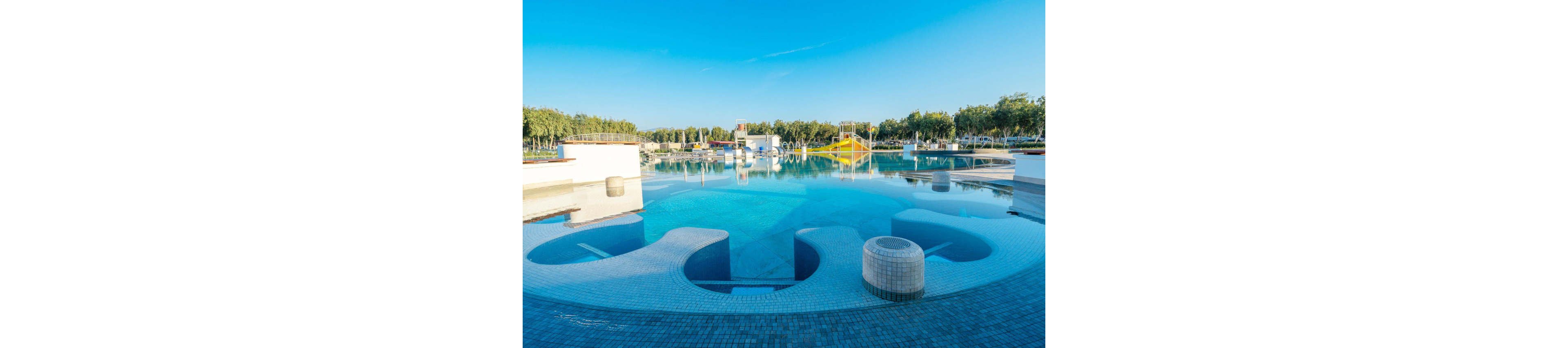A large outdoor swimming pool features curved, tiled seating areas and shallow lounging zones. The water is calm and reflects the bright blue sky above. In the background, additional pool structures and play areas are visible, surrounded by trees and open space. The scene suggests a modern aquatic center or resort on a sunny day.