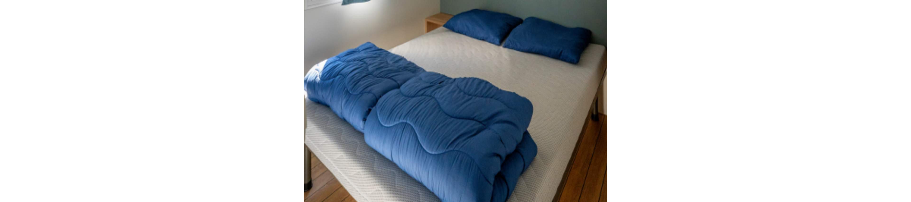 A neatly folded blue comforter lies across the foot of an unmade bed. Two matching blue pillows rest at the head of the bed on a light-colored mattress. The room features wooden flooring and simple, minimal furniture. Soft natural light enters from the side, creating a calm and cozy bedroom atmosphere.