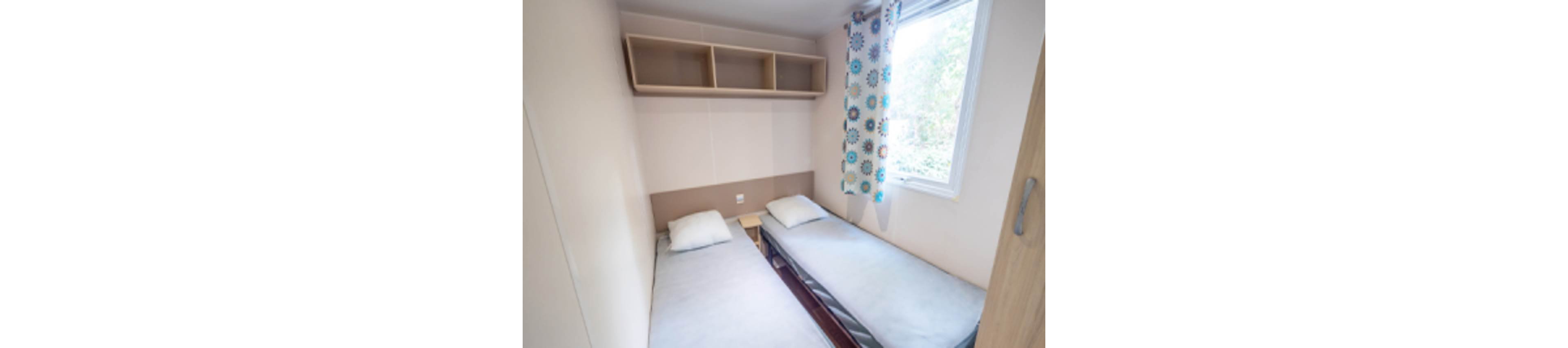 Compact bedroom featuring two single beds placed parallel along the walls. A large window with patterned curtains lets in natural light and offers a view of greenery outside. Minimalist wooden shelving is mounted above one bed for storage. The room has a clean, simple design suitable for a guest room or vacation rental.