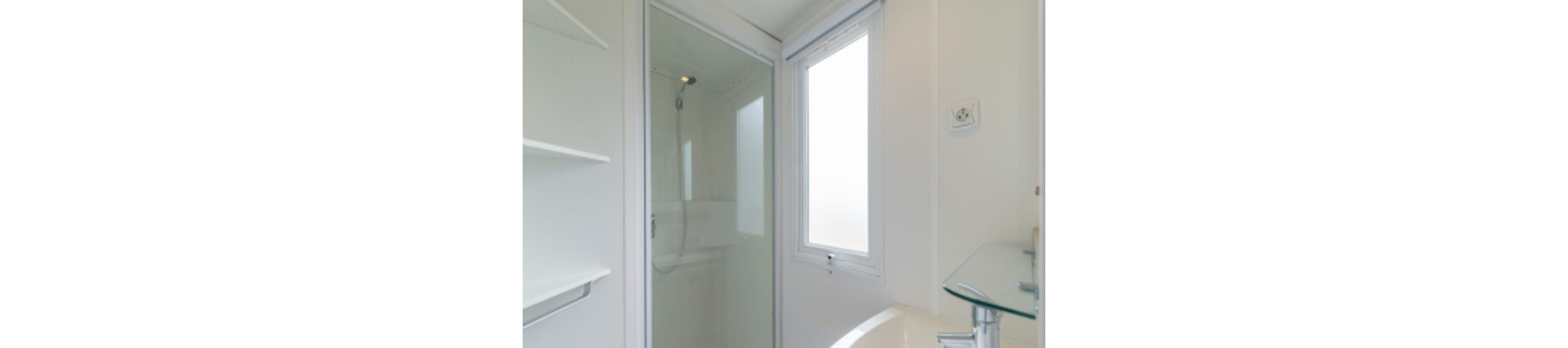 A compact modern bathroom features a glass-enclosed shower with a handheld showerhead. A frosted window lets in natural light while maintaining privacy. White walls, shelving, and fixtures create a clean, minimalist look. A sleek glass sink countertop and chrome faucet add a contemporary touch to the small space.