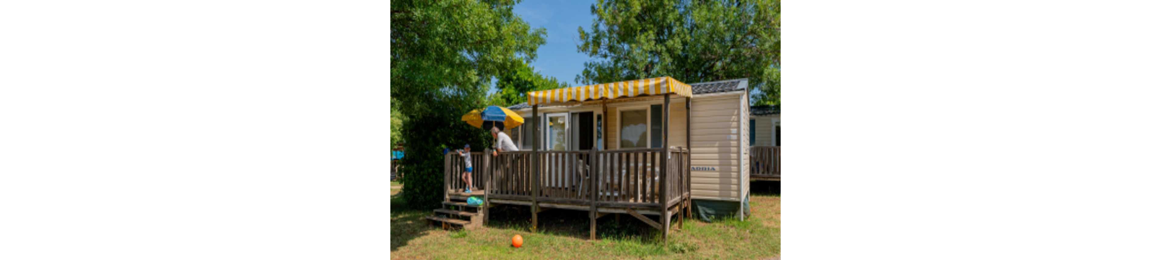 A small mobile home with a wooden terrace stands in a grassy, tree-filled campsite. A striped yellow and white awning shades the deck where an adult and child are spending time together. Another child stands on the steps near a colorful umbrella, and a bright orange ball lies on the grass. The scene suggests a relaxed summer holiday in a natural outdoor setting.