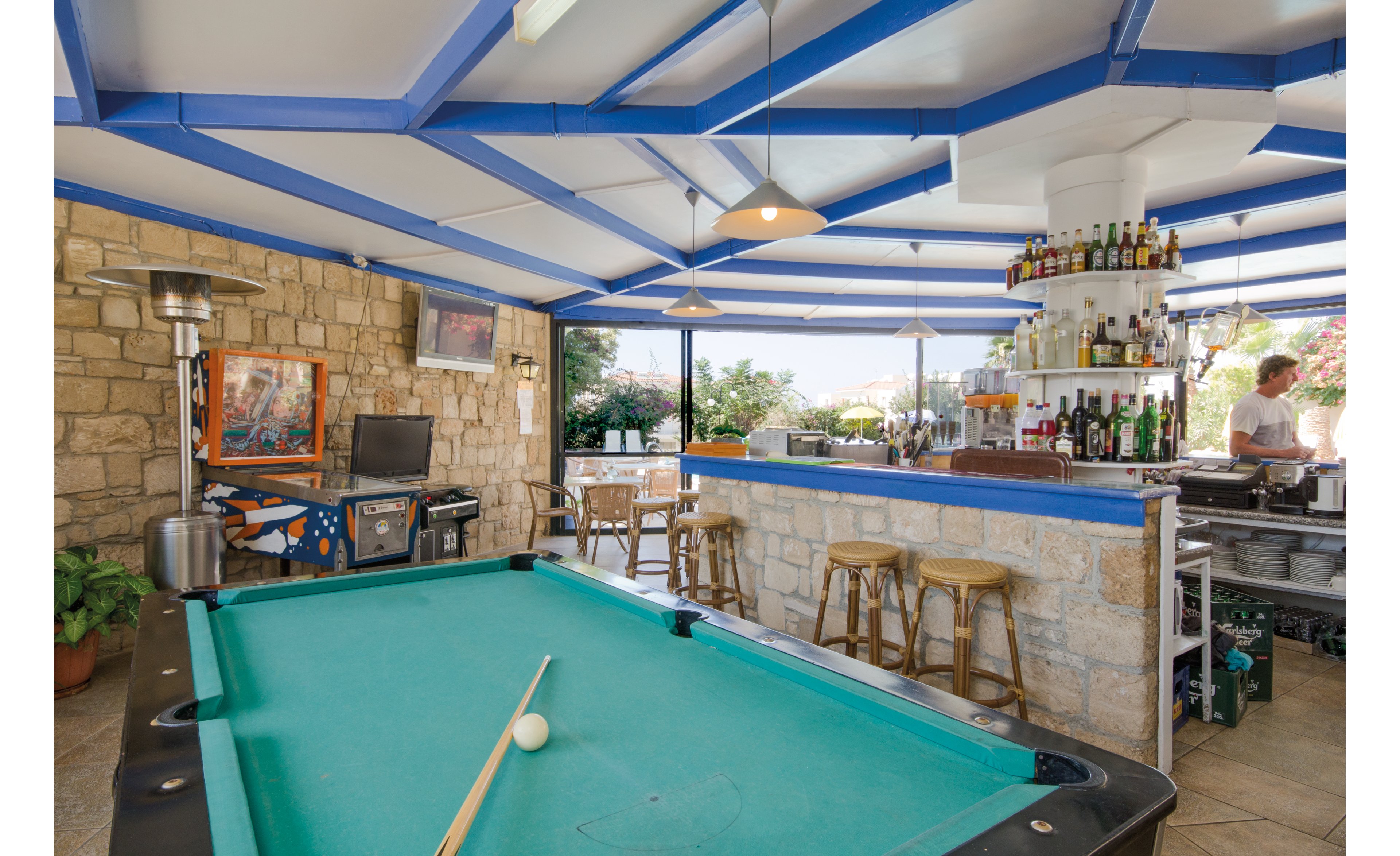 A pool table with a cue and ball is the focal point in a lively bar area. The setting features stone walls, blue accents, and a well-stocked bar with stools. A pinball machine and television are visible, adding to the recreational atmosphere. The space is bright and open, with natural light streaming in from large windows.