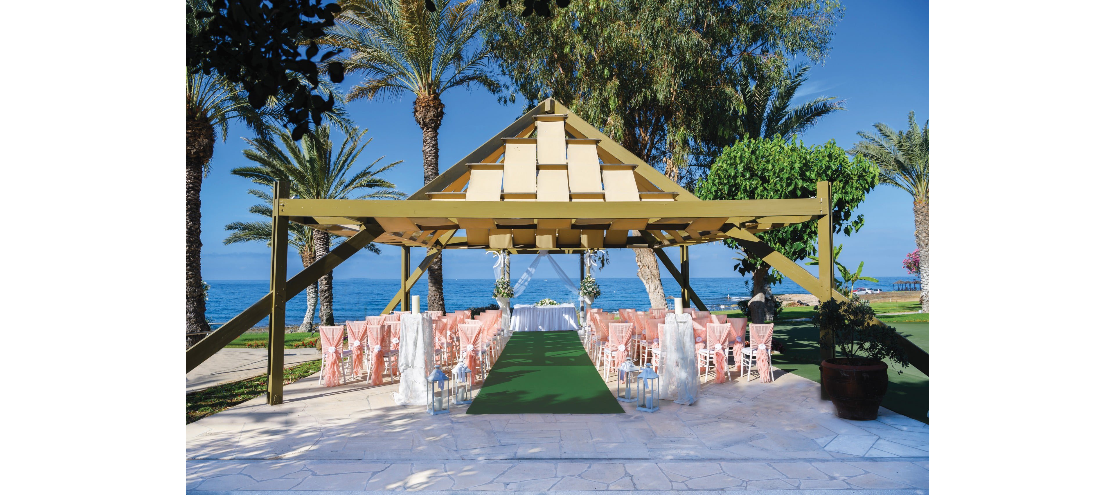 An elegant outdoor wedding ceremony is arranged beneath a wooden pavilion by the sea. Rows of pink chairs line a green aisle runner leading to a white altar table decorated with flowers. Tall palm trees and lush greenery frame the scene against a bright blue sky and calm ocean. The setting suggests a tropical or Mediterranean destination wedding venue.