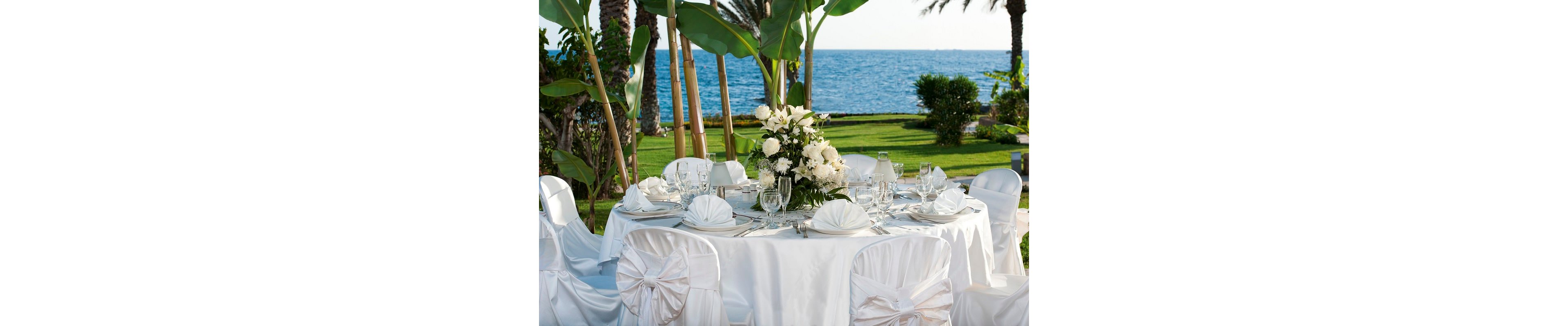 A round table is beautifully set for a formal event with a view of the ocean in the background. White linens, folded napkins, and chair covers create a refined and cohesive look. A large floral centerpiece with white flowers and greenery stands in the middle of the table, surrounded by glassware and tableware. Palm trees, lush grass, and tropical plants frame the seaside setting, suggesting a romantic wedding or special celebration.