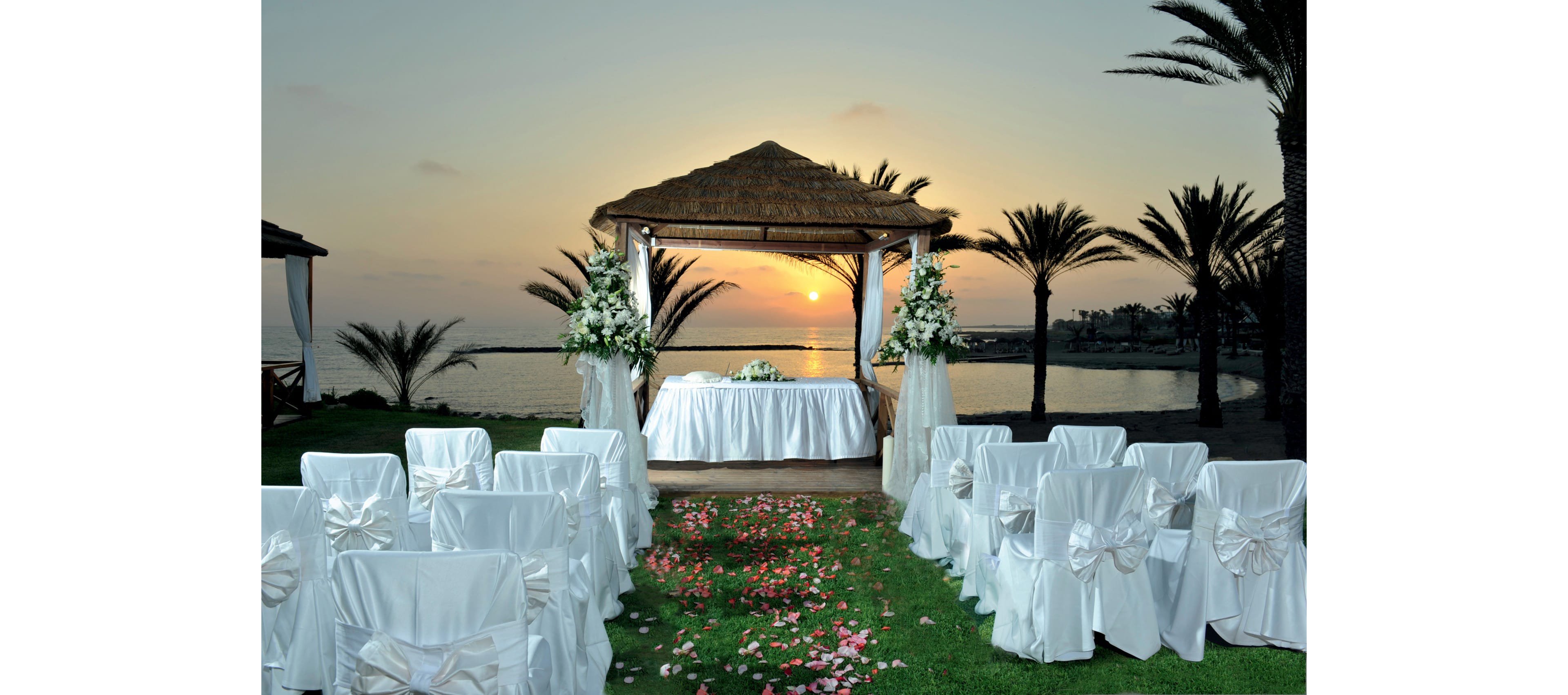 An outdoor wedding ceremony setup is arranged under a thatched gazebo by the sea at sunset. White chairs with large ribbon bows face a flower-adorned altar table on a manicured lawn. Palm trees frame the scene while the sun sets over calm water in the background. Pink flower petals are scattered along the aisle, adding a romantic touch to the elegant white decor.